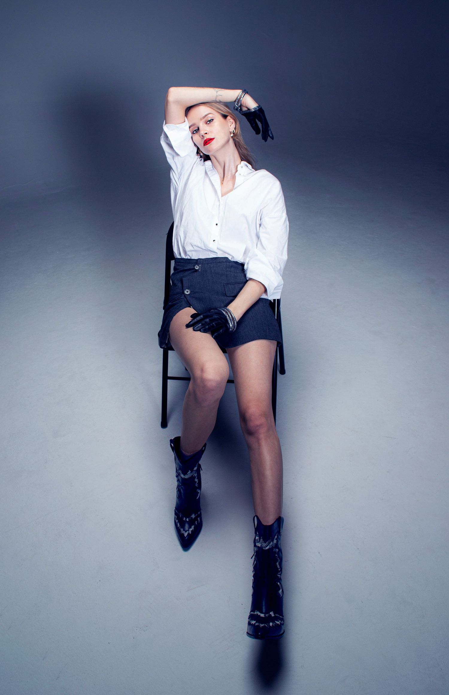 Woman in white shirt, dark shorts, and cowboy boots posing on a chair in a studio.