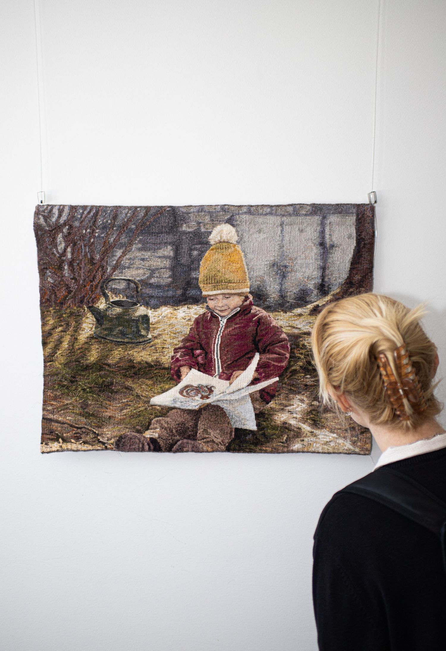 Woven tapestry of child in yellow hat reading book outdoors displayed on white gallery wall.