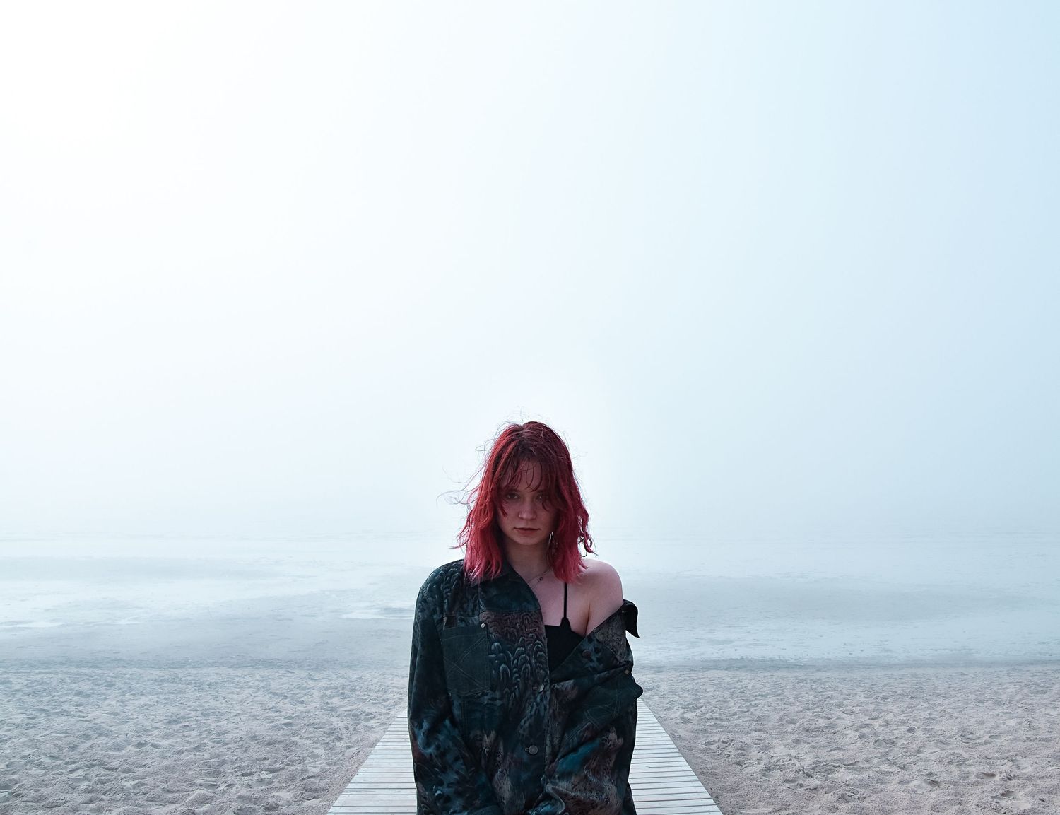 Red-haired woman wrapped in dark fabric standing on a foggy beach boardwalk by the sea.