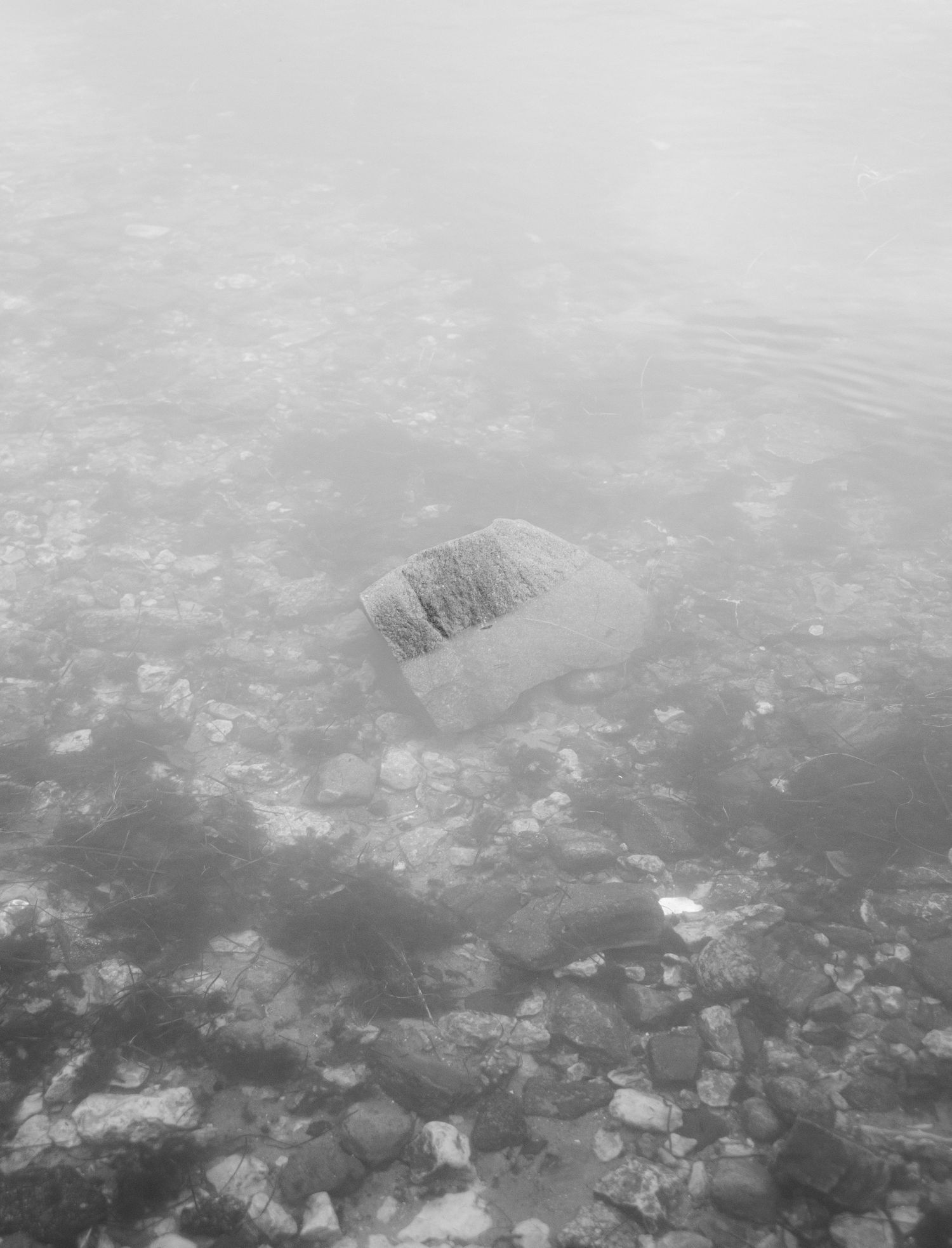 Black and white aerial view of a single rock submerged in shallow clear water with pebbles visible on the bottom.