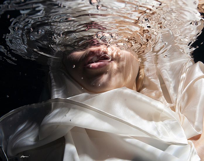 Underwater portrait of person submerged in water wearing white fabric with rippling water surface above.