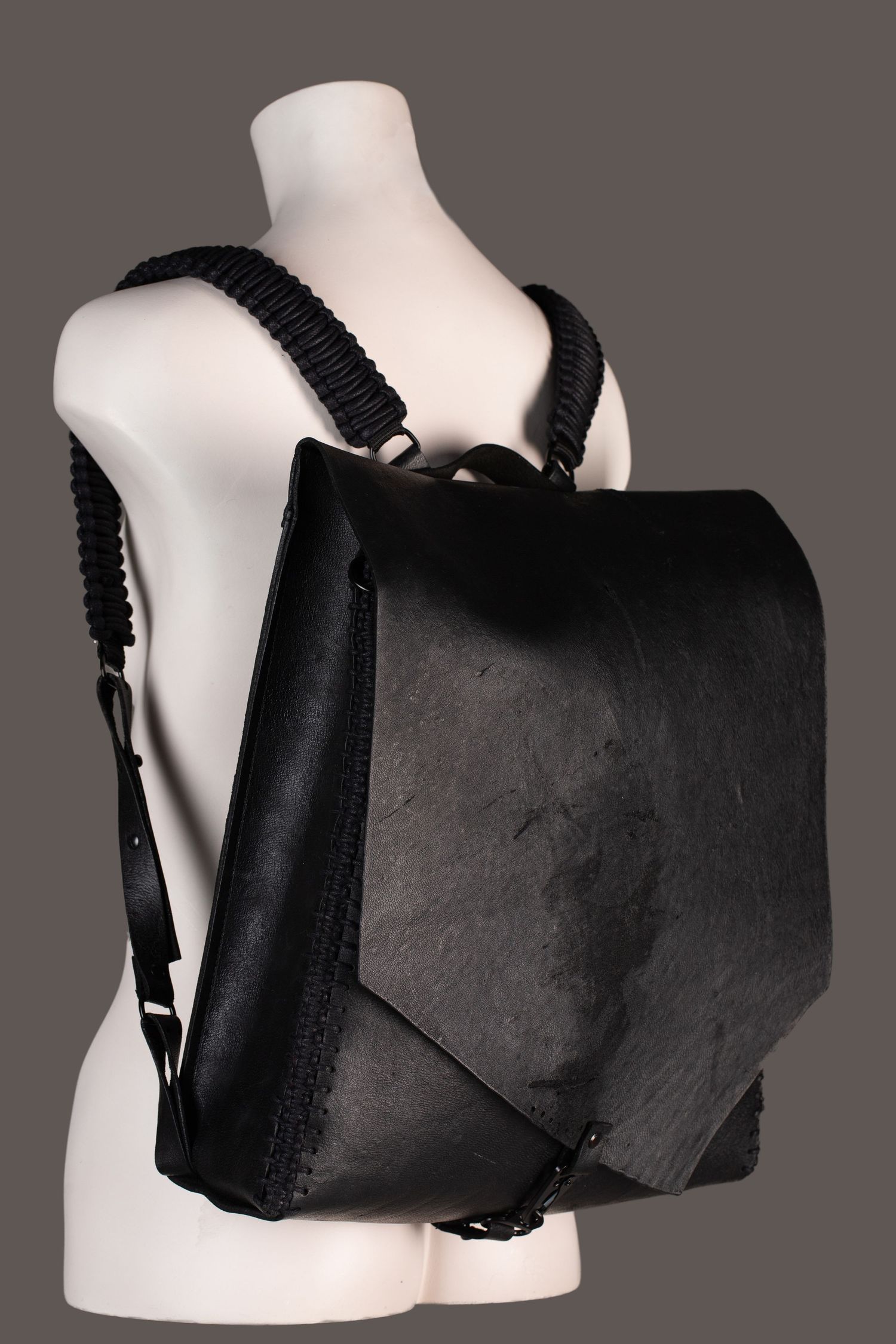 Black leather backpack with braided straps displayed on a white mannequin against a gray background.