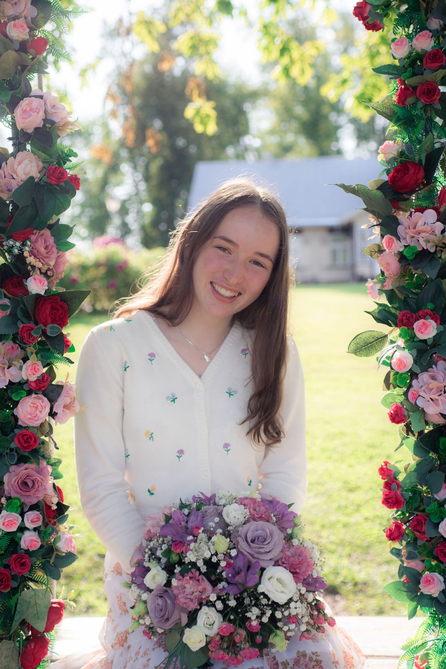 Smiling young woman in white cardigan holding a bouquet of flowers under a floral arch outdoors.