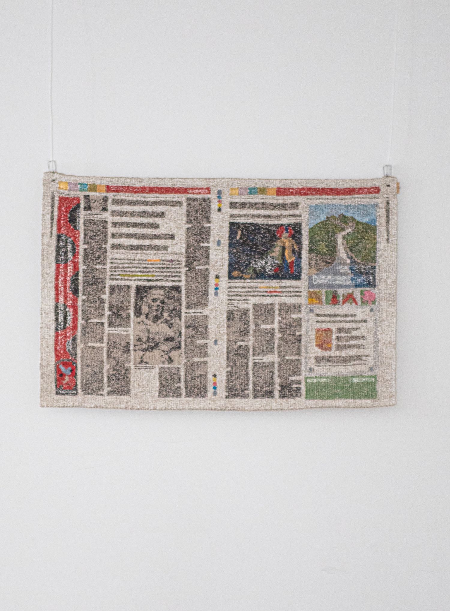 Detailed embroidered textile artwork replicating a newspaper spread hanging on white wall.