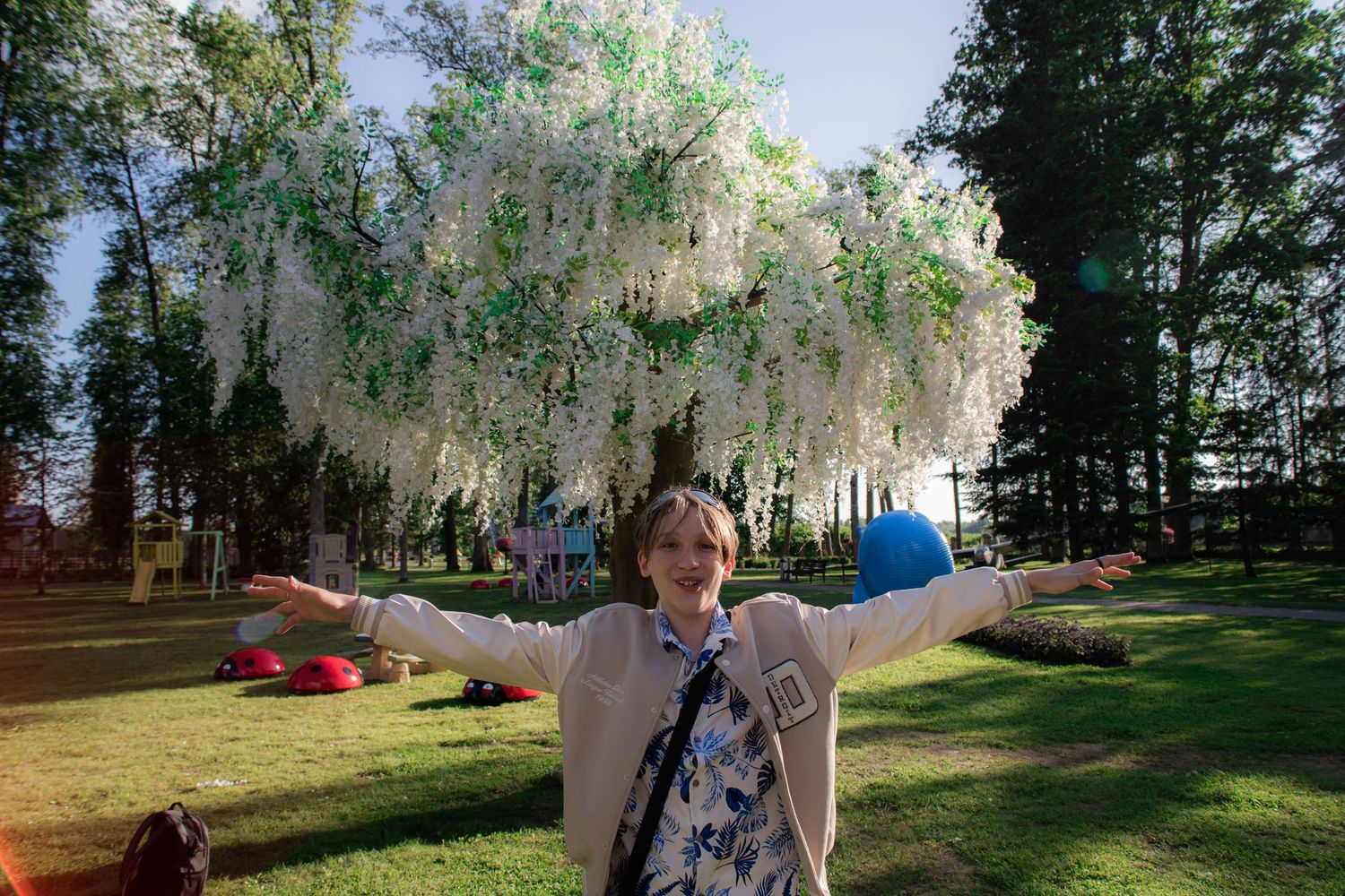 Joyful woman posing with arms outstretched under white wisteria flowers in a vibrant park.