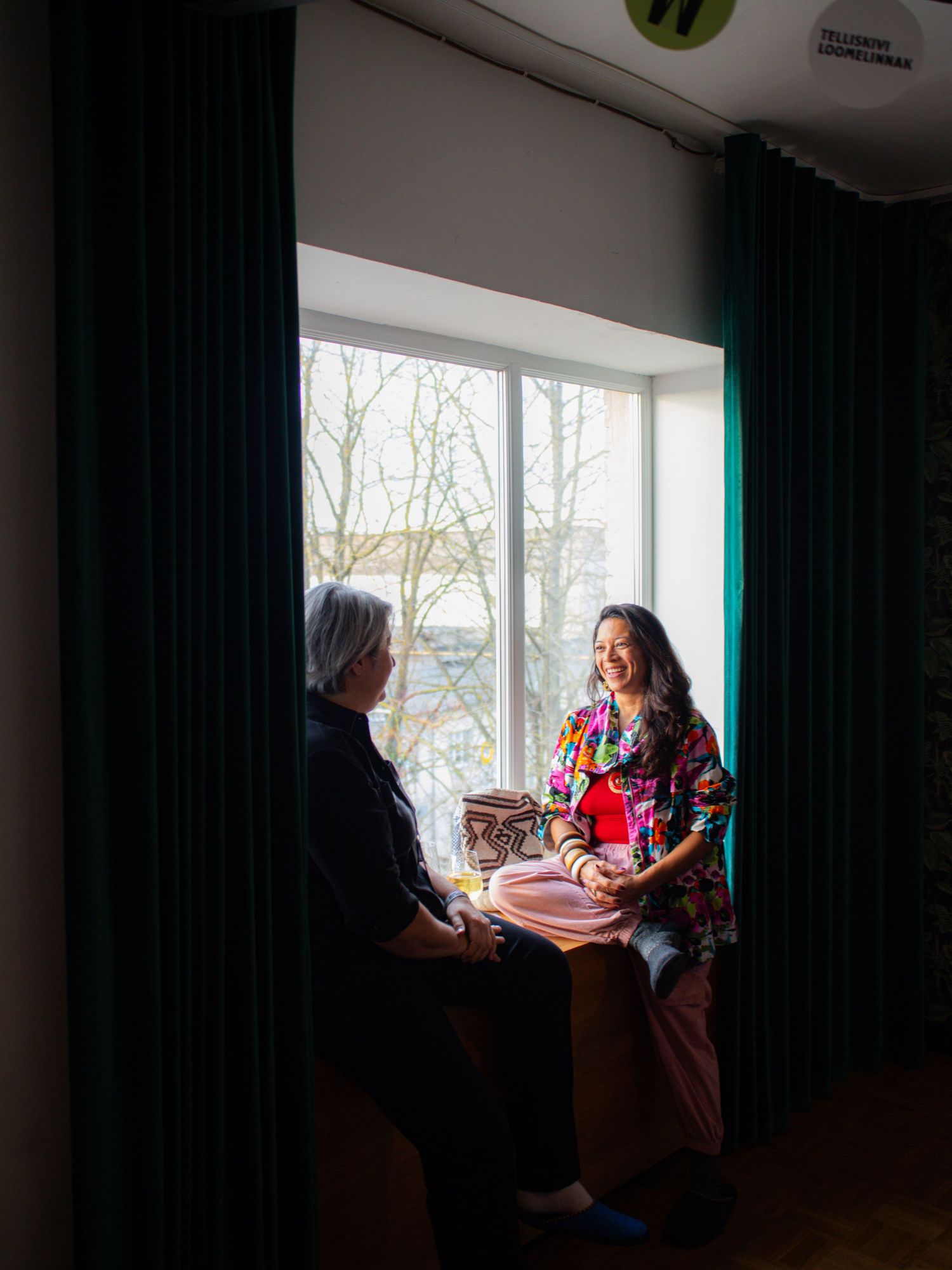 Two women having a conversation by a large window framed by dark teal curtains in a bright room.