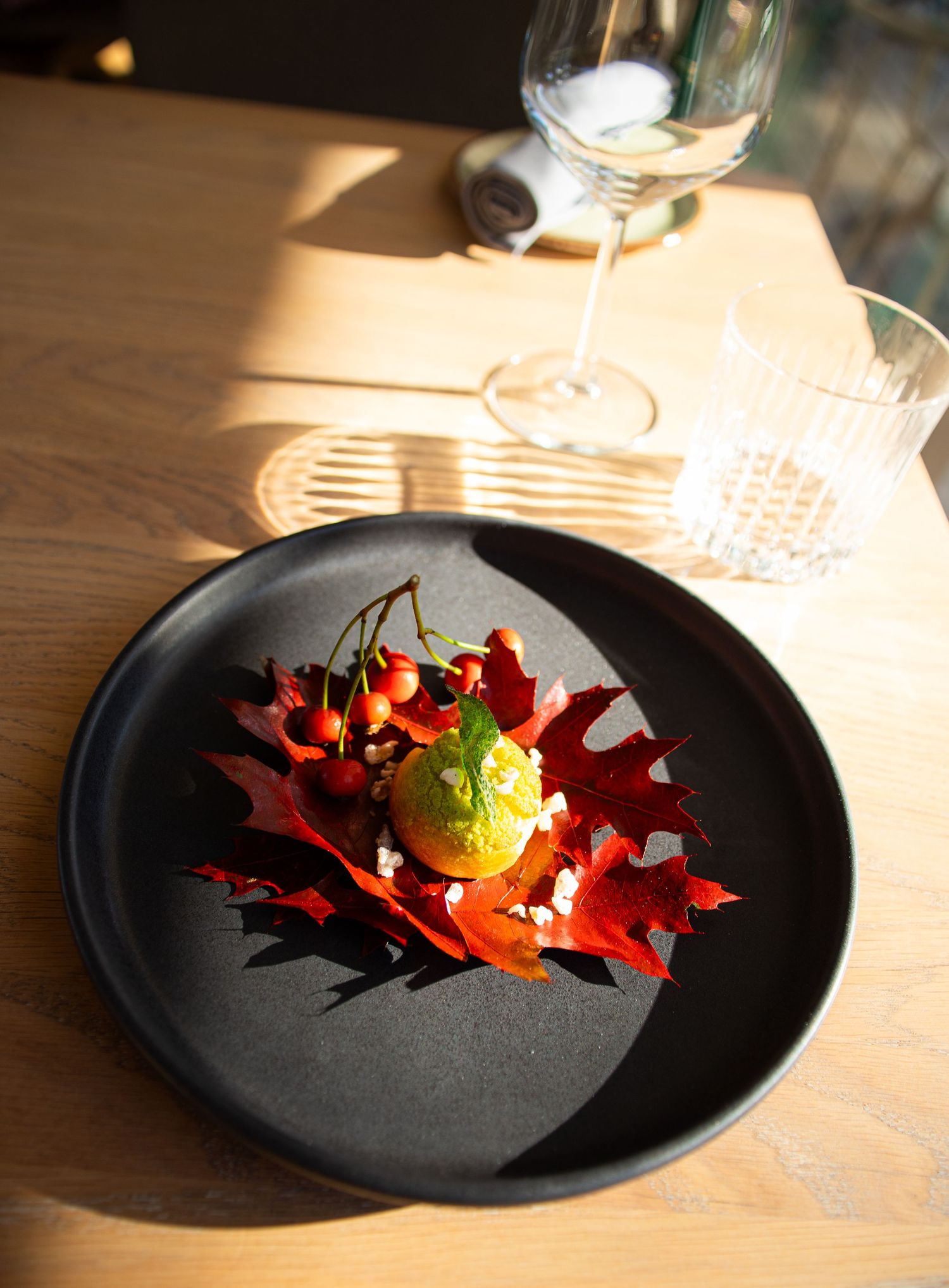 Artistic autumn dish with red maple leaves, orange citrus, green herb sorbet, and berries on a black plate with wine glass.