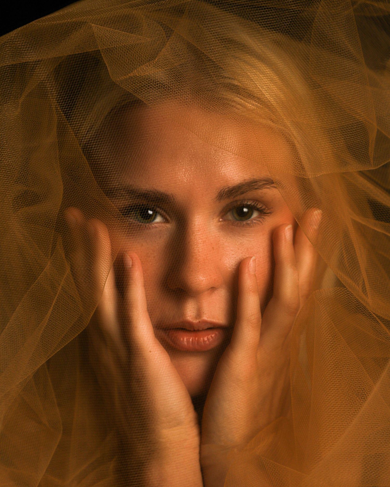 Blonde woman with freckles gazing forward, face cradled in hands, draped in golden tulle veil.