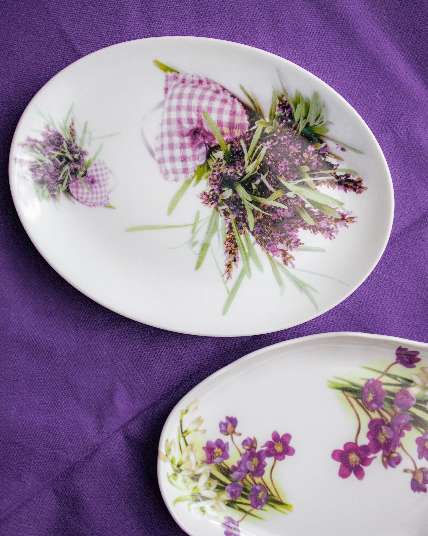 Two white oval porcelain plates with purple lavender floral designs on a violet fabric background.