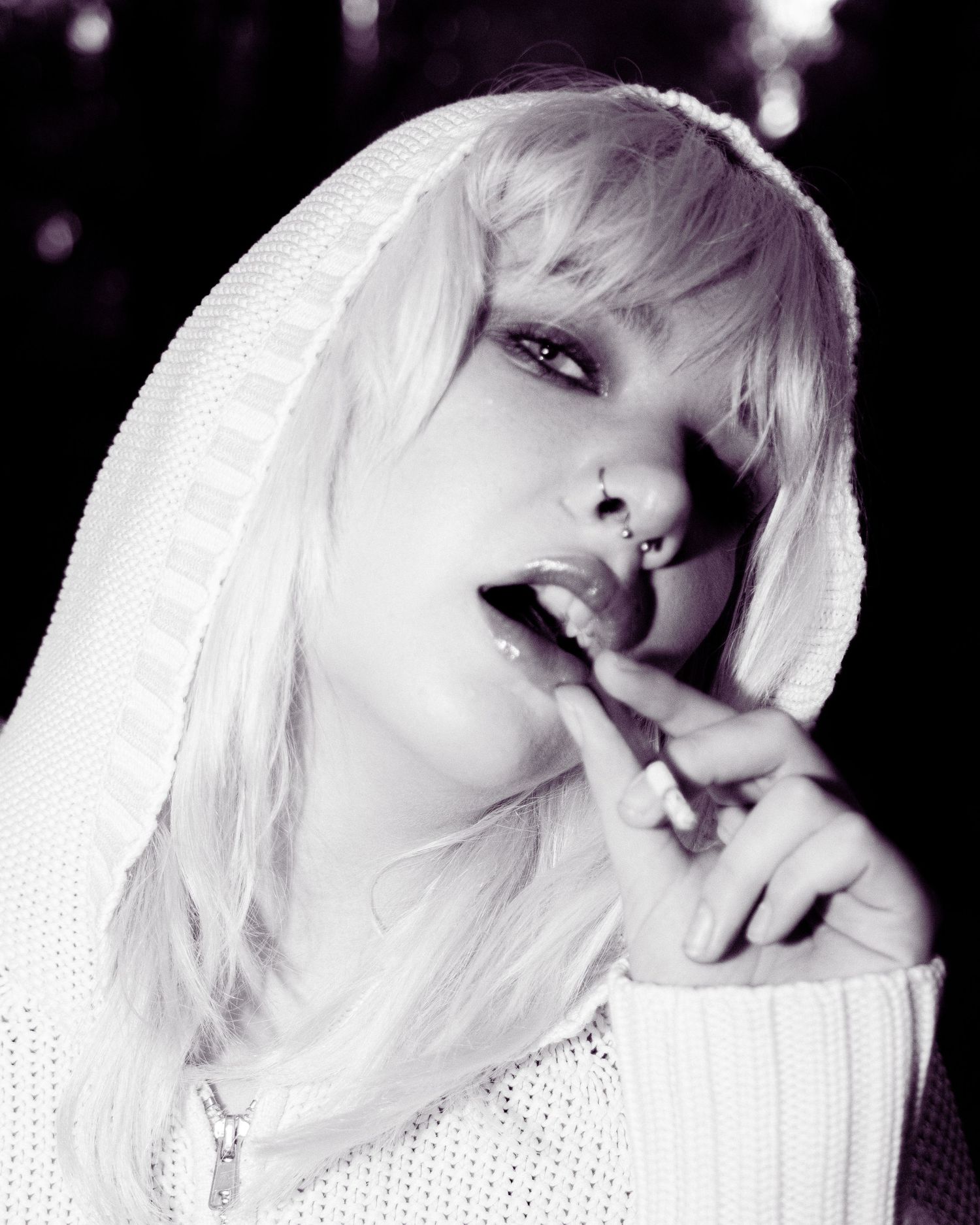Black and white portrait of platinum blonde woman in white hoodie with lip piercing licking finger.