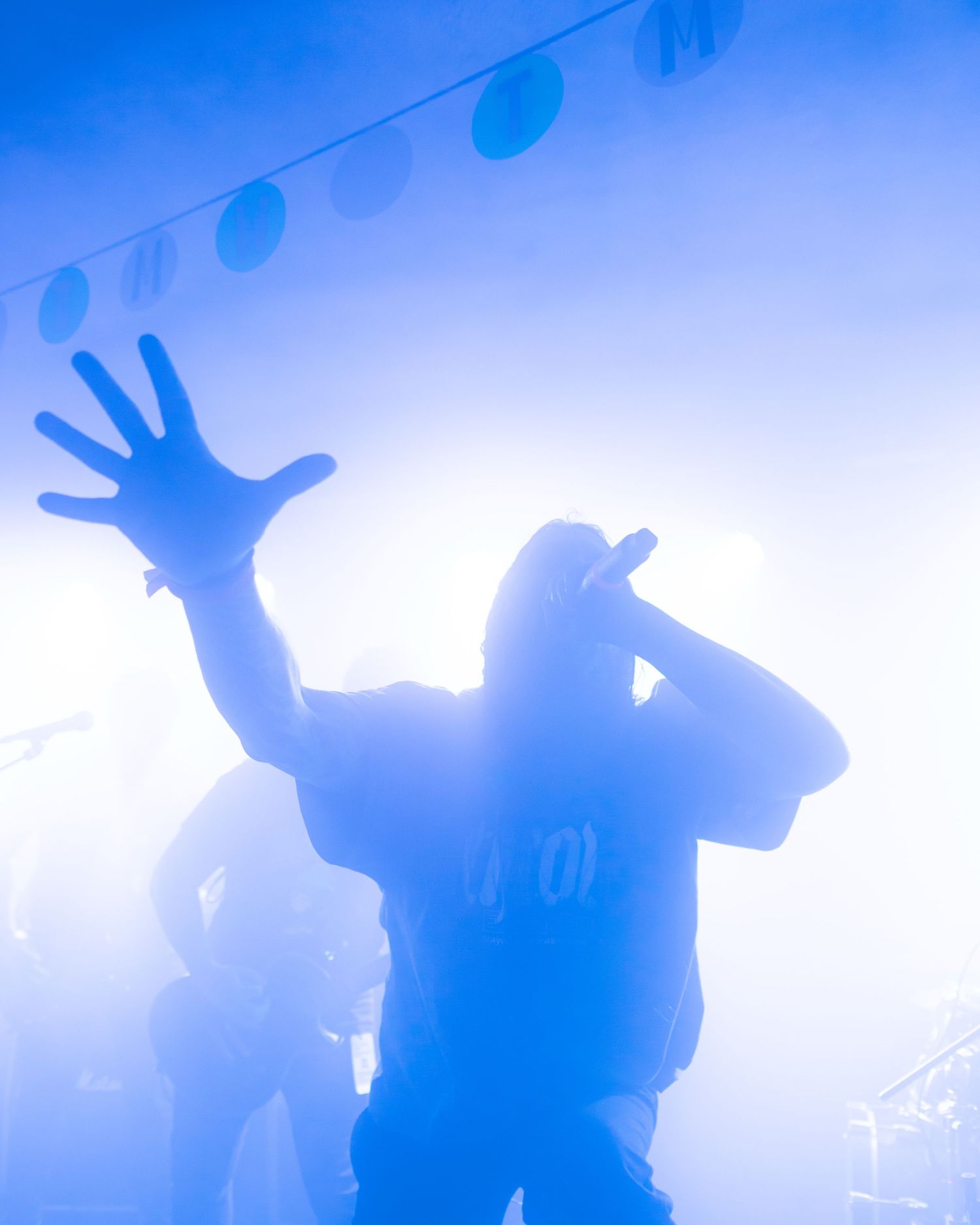 Silhouetted performer singing into microphone with hand raised on fog-filled stage bathed in blue concert lighting.