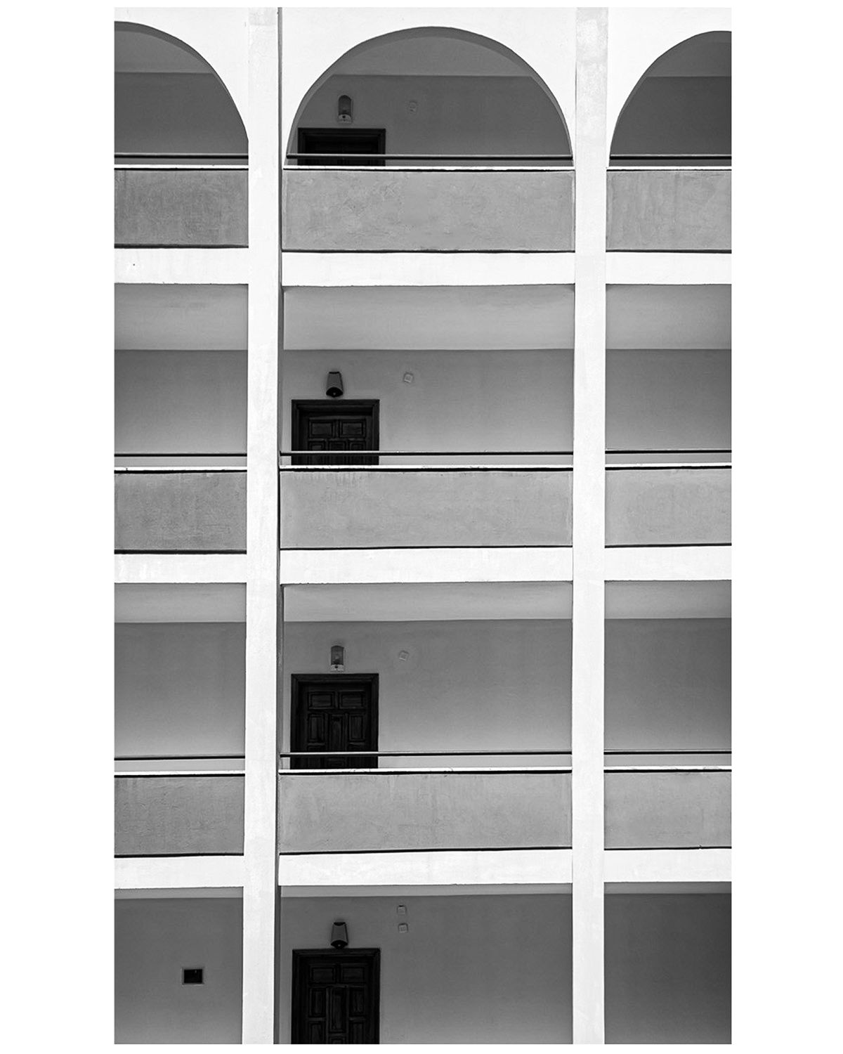 Black and white architectural photo of a multi-story building facade with arched windows and repeating grid pattern.