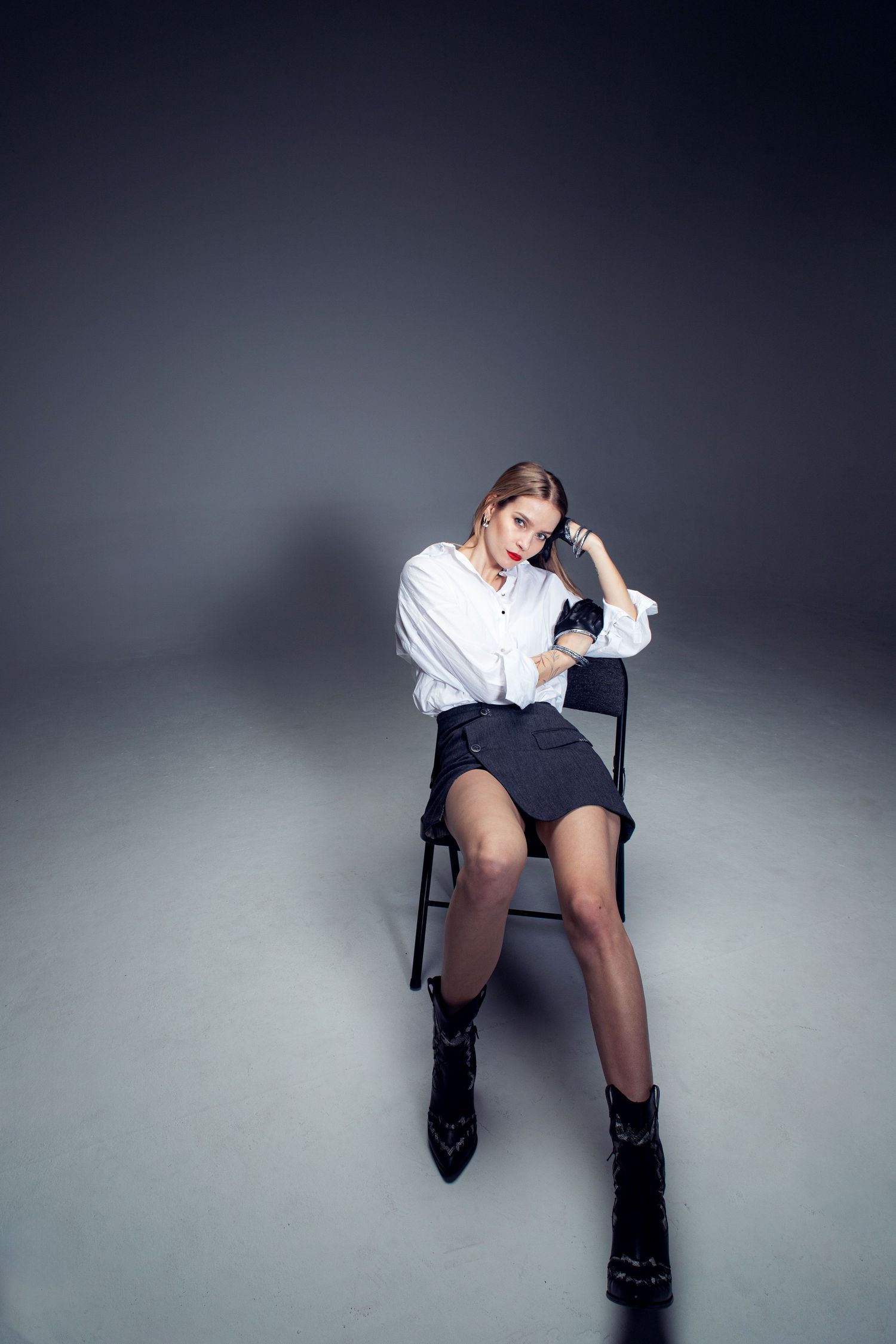 Woman in white shirt and dark mini skirt poses on chair with black ankle boots in studio.