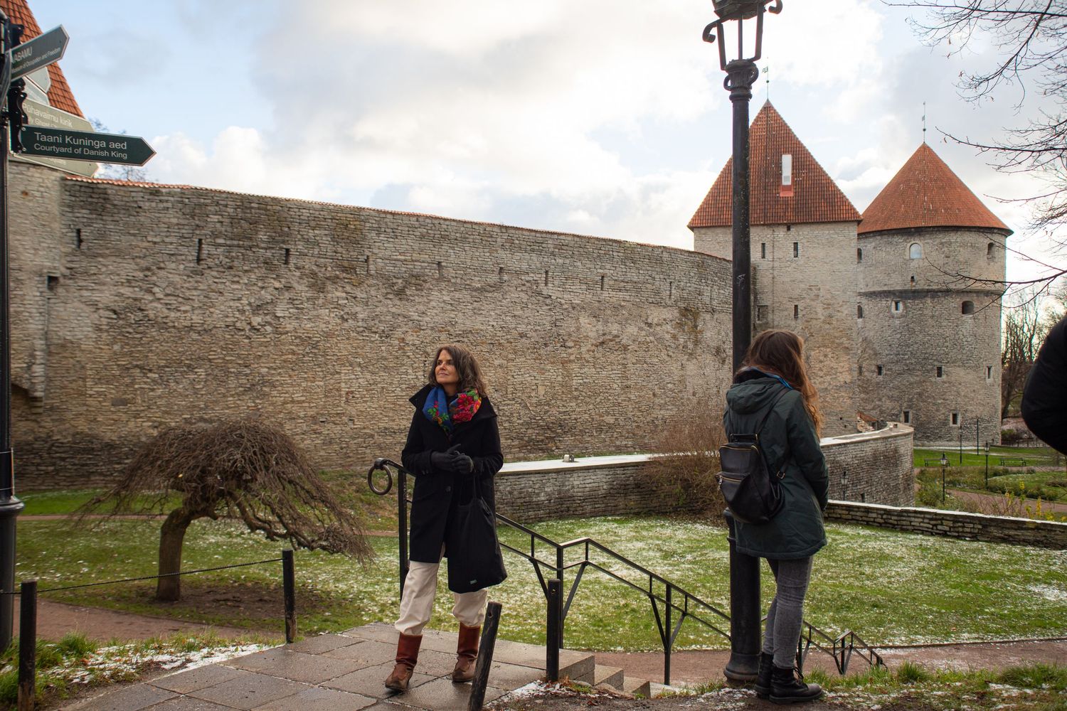Two women stand near a historic stone city wall with towers in a European old town.