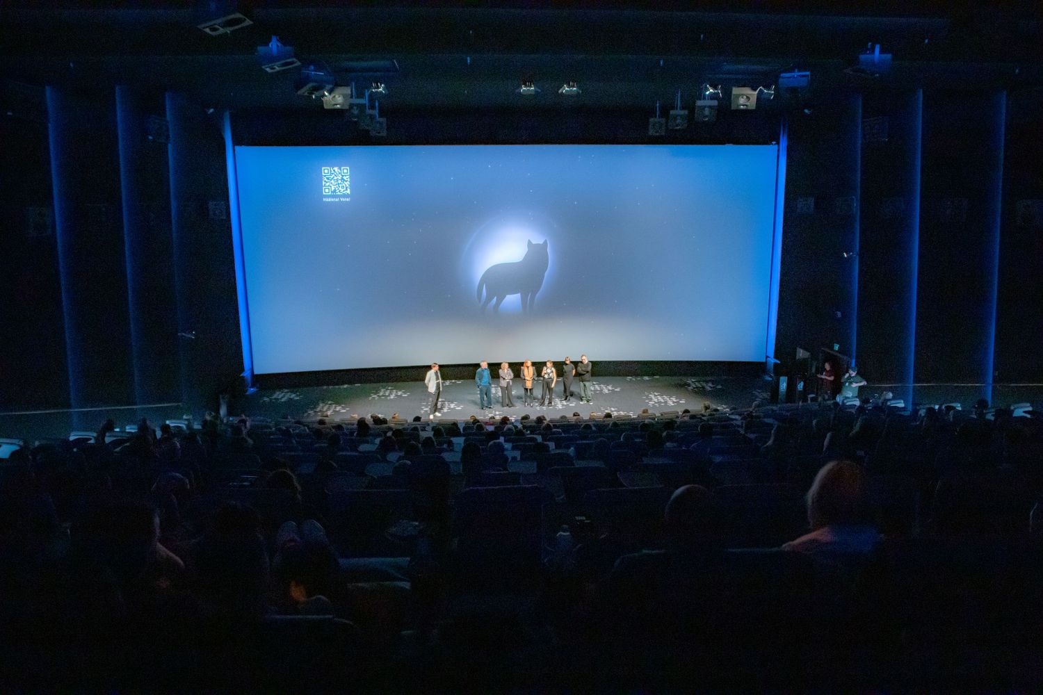 A wide-angle view of a packed cinema audience watching presenters on stage beneath a large illuminated screen.