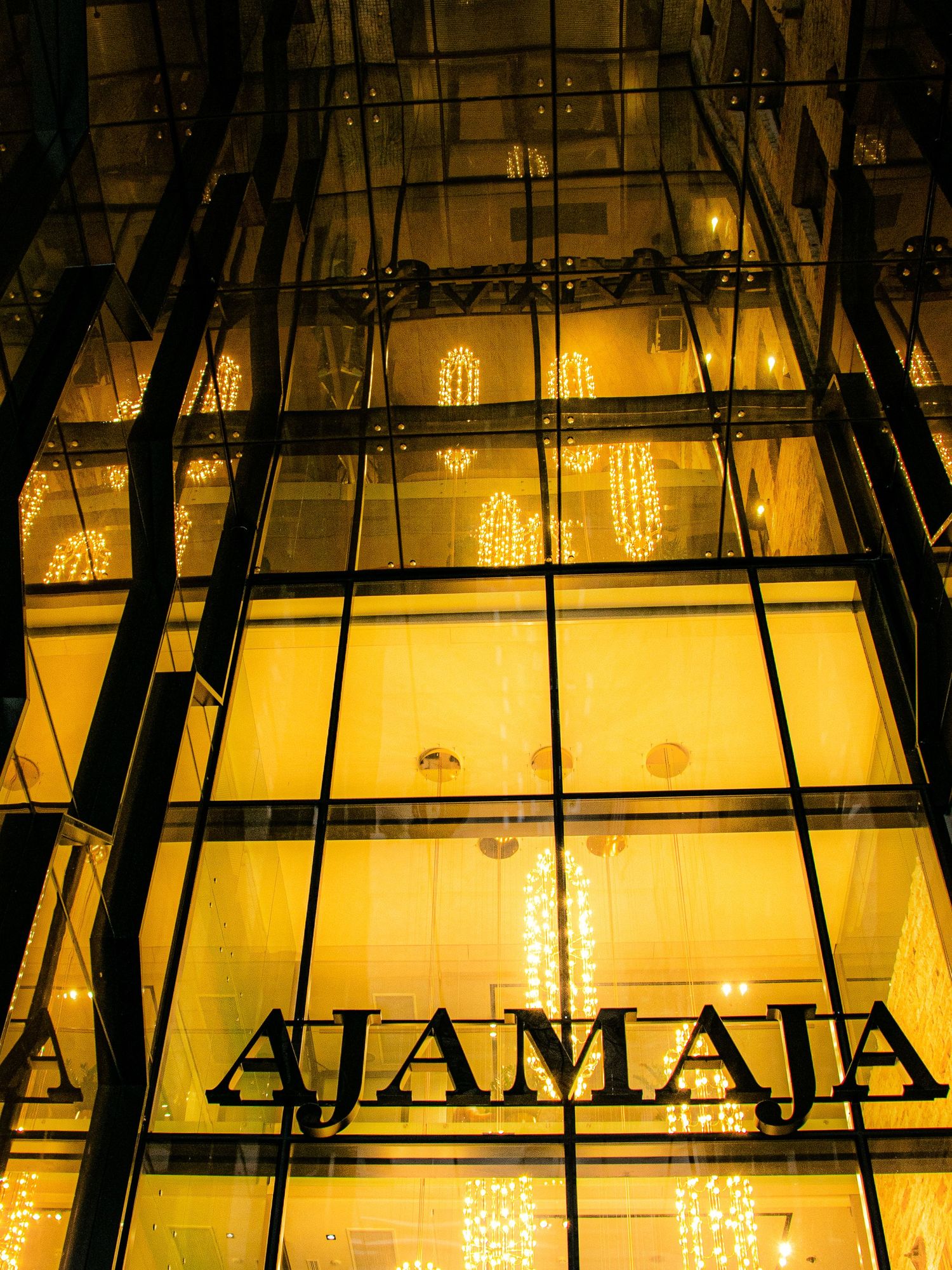 Illuminated golden glass facade of Ajamaja building at night with warm chandelier reflections and bold signage.