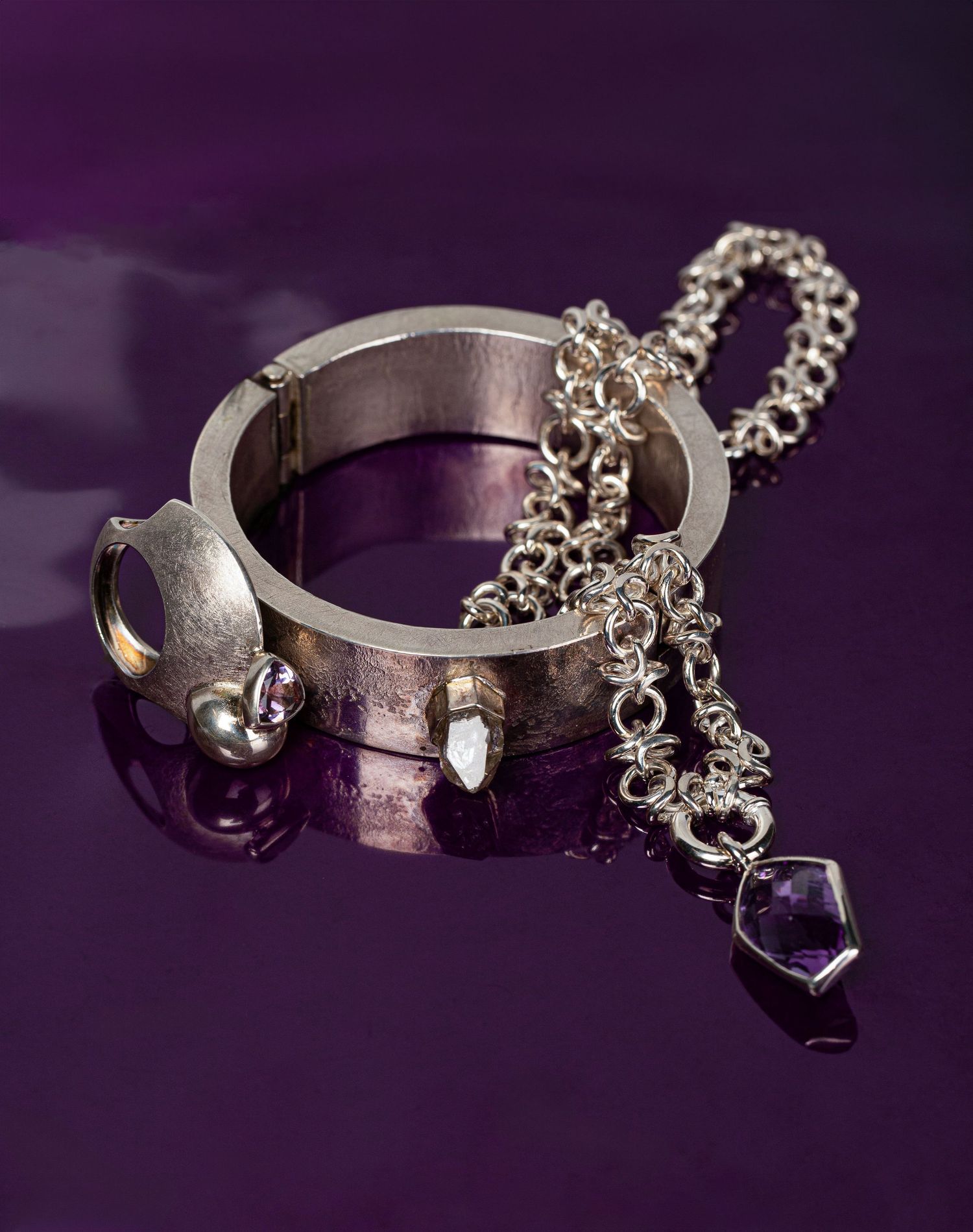 Unique silver cuff bracelet with chain and amethyst crystal pendant on reflective purple background.