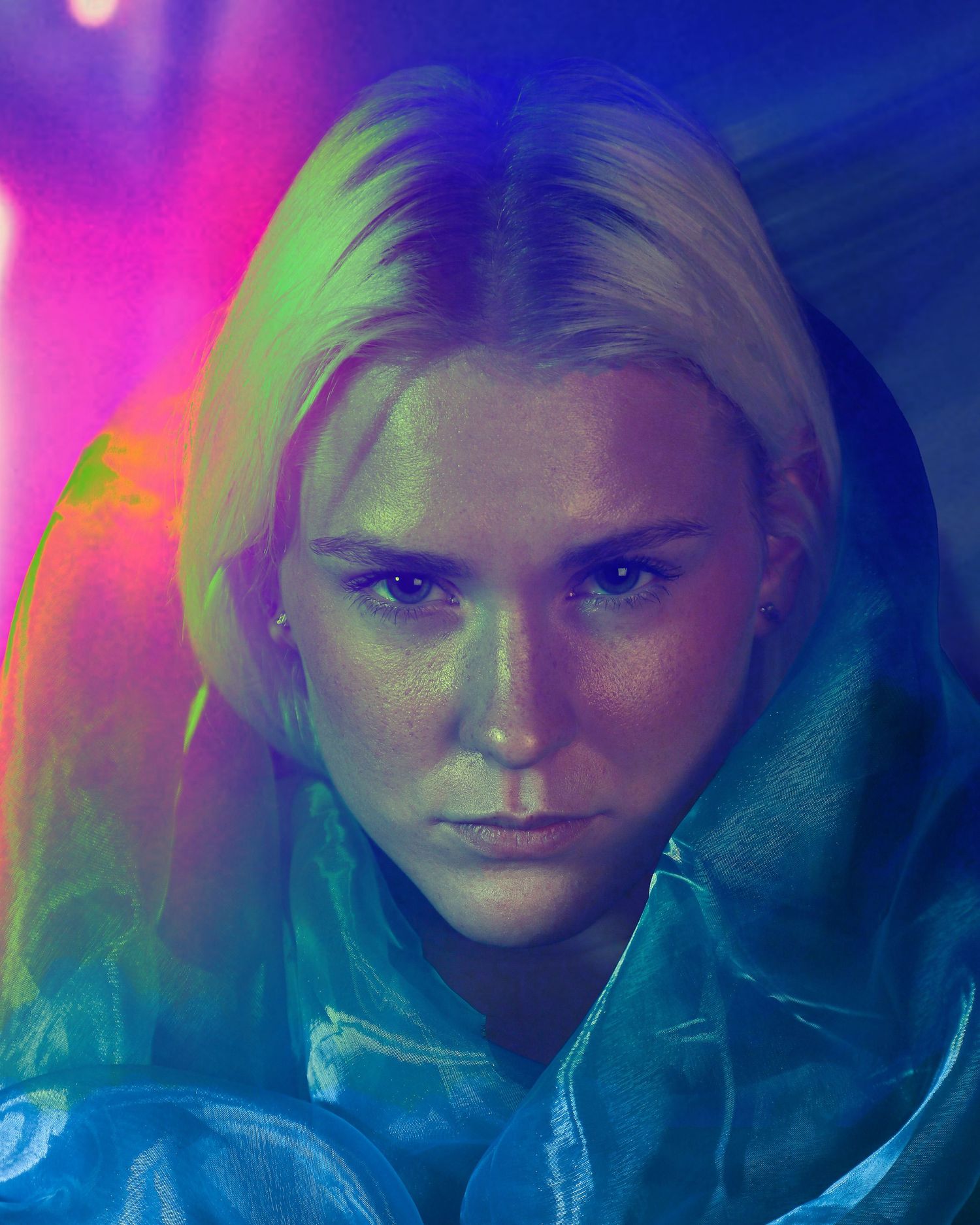 A blonde woman bathed in vibrant neon pink and green lights with an intense gaze.
