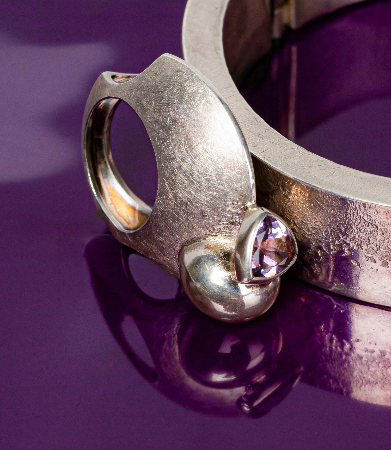 Silver ring with amethyst gemstone accent displayed on purple reflective surface.