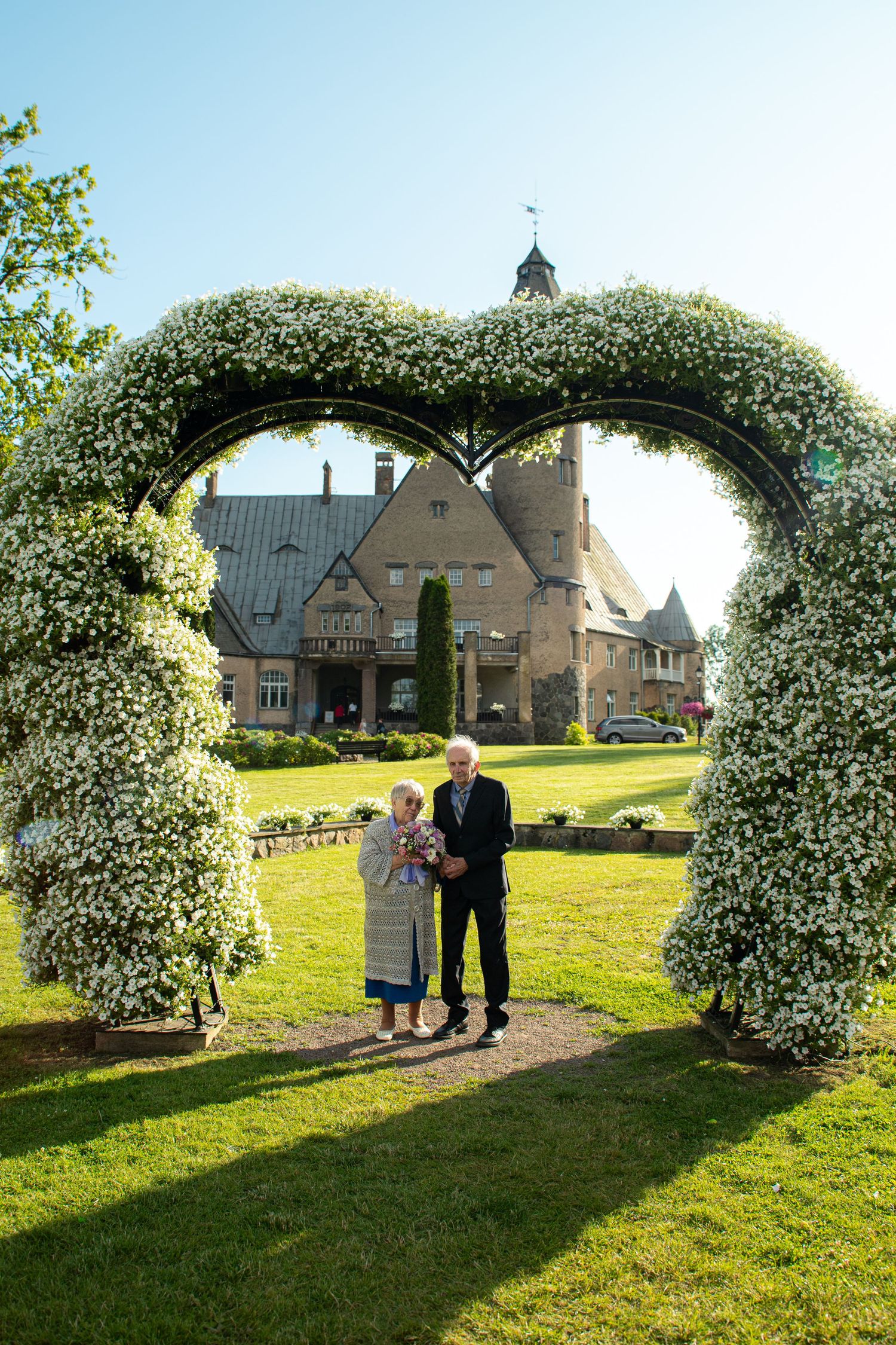 Elderly couple standing under a heart-shaped floral arch with a historic stone manor in background.