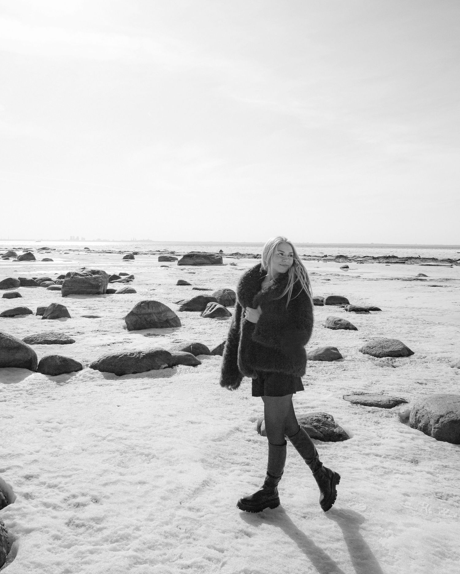 Black and white photo of a young woman in a fur coat walking on a rocky, snow-covered beach in winter.