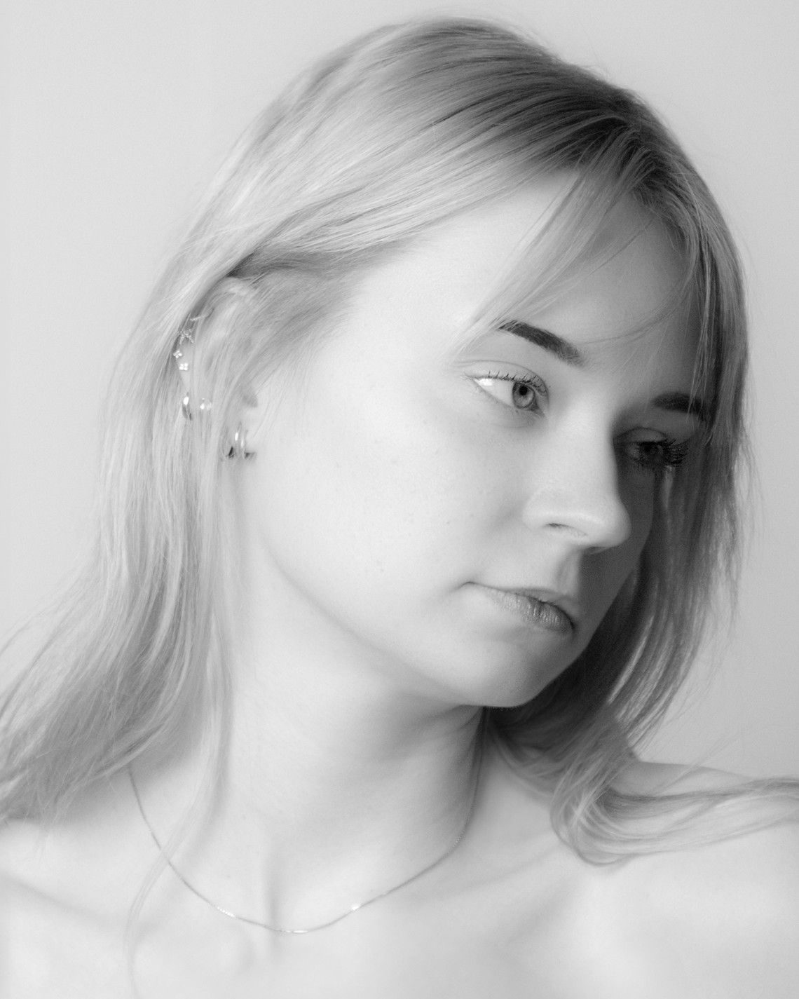 Black and white portrait of a young woman with blonde hair wearing delicate ear pins and a thin chain necklace.