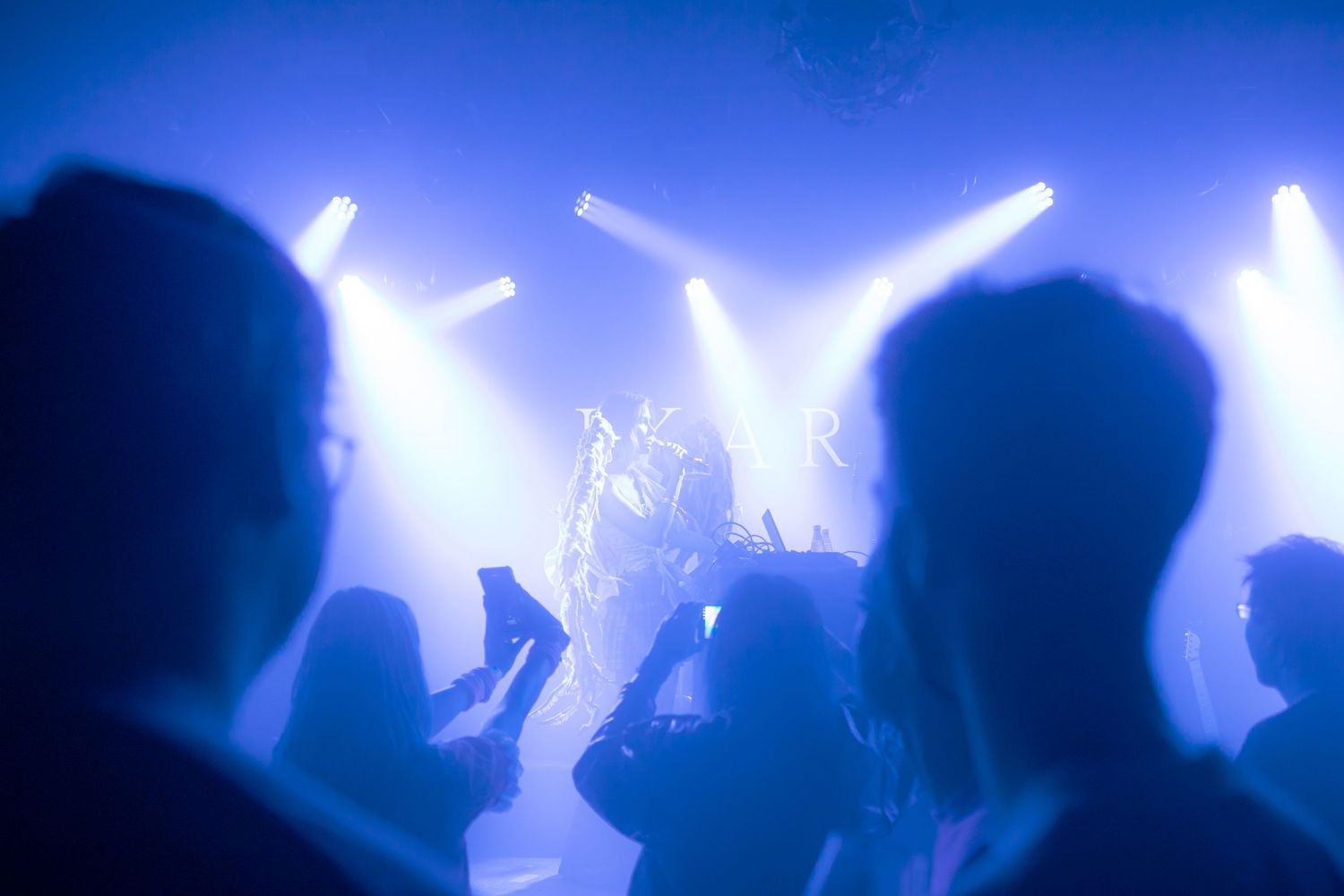 Crowd of silhouetted concert-goers bathed in blue stage lights watching a live performance with bright spotlights.