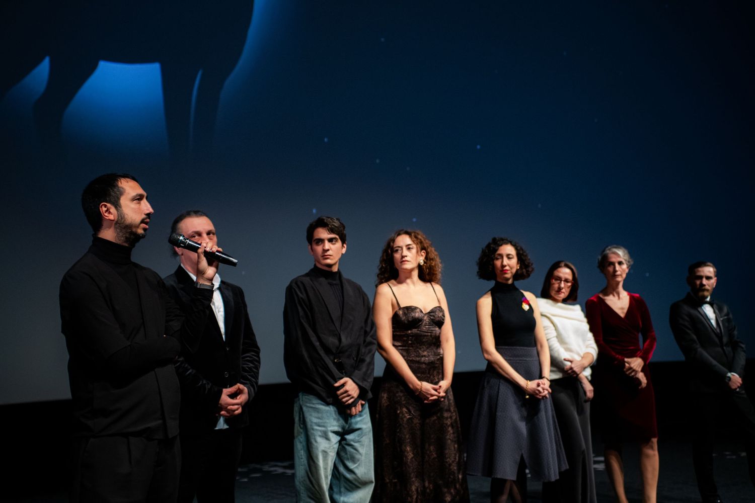 Film cast and crew standing on stage at a movie premiere under dramatic blue lighting.