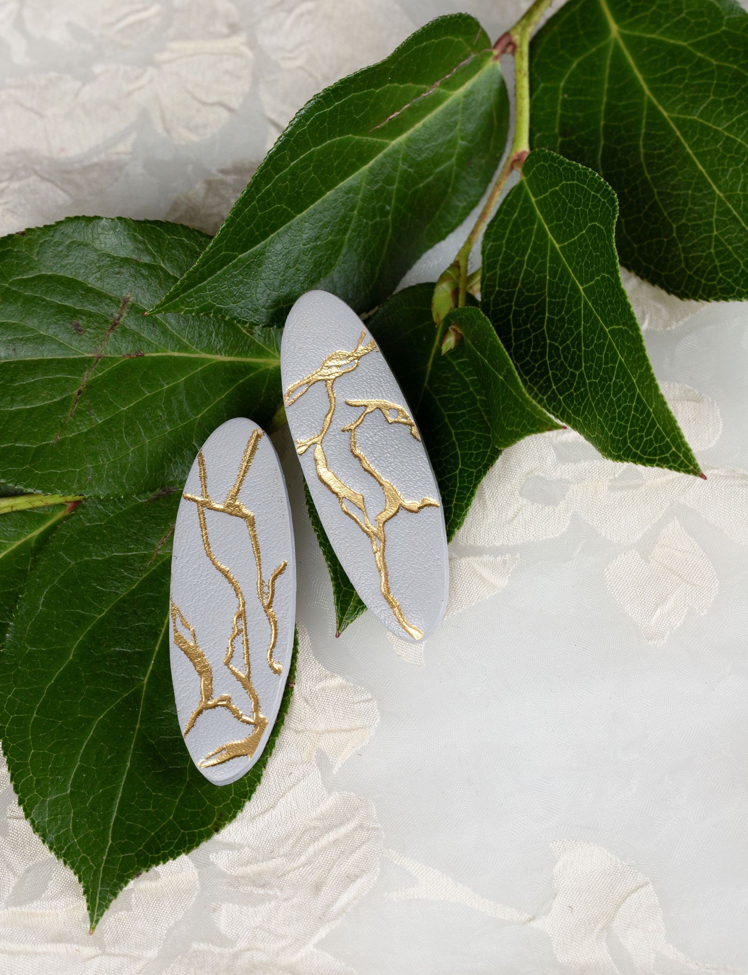 White oval earrings with gold kintsugi-style crack pattern resting on green leaves.