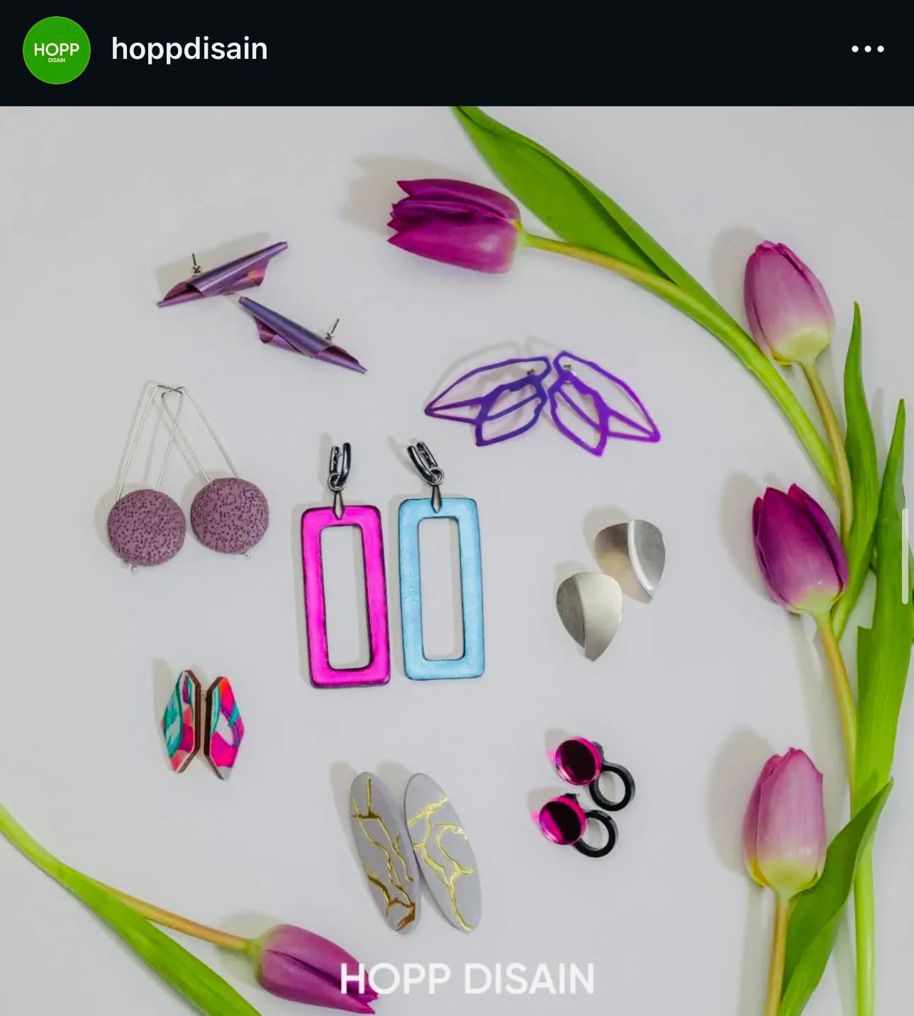 Colorful collection of modern geometric earrings by Hopp Disain arranged with purple tulips on white background.