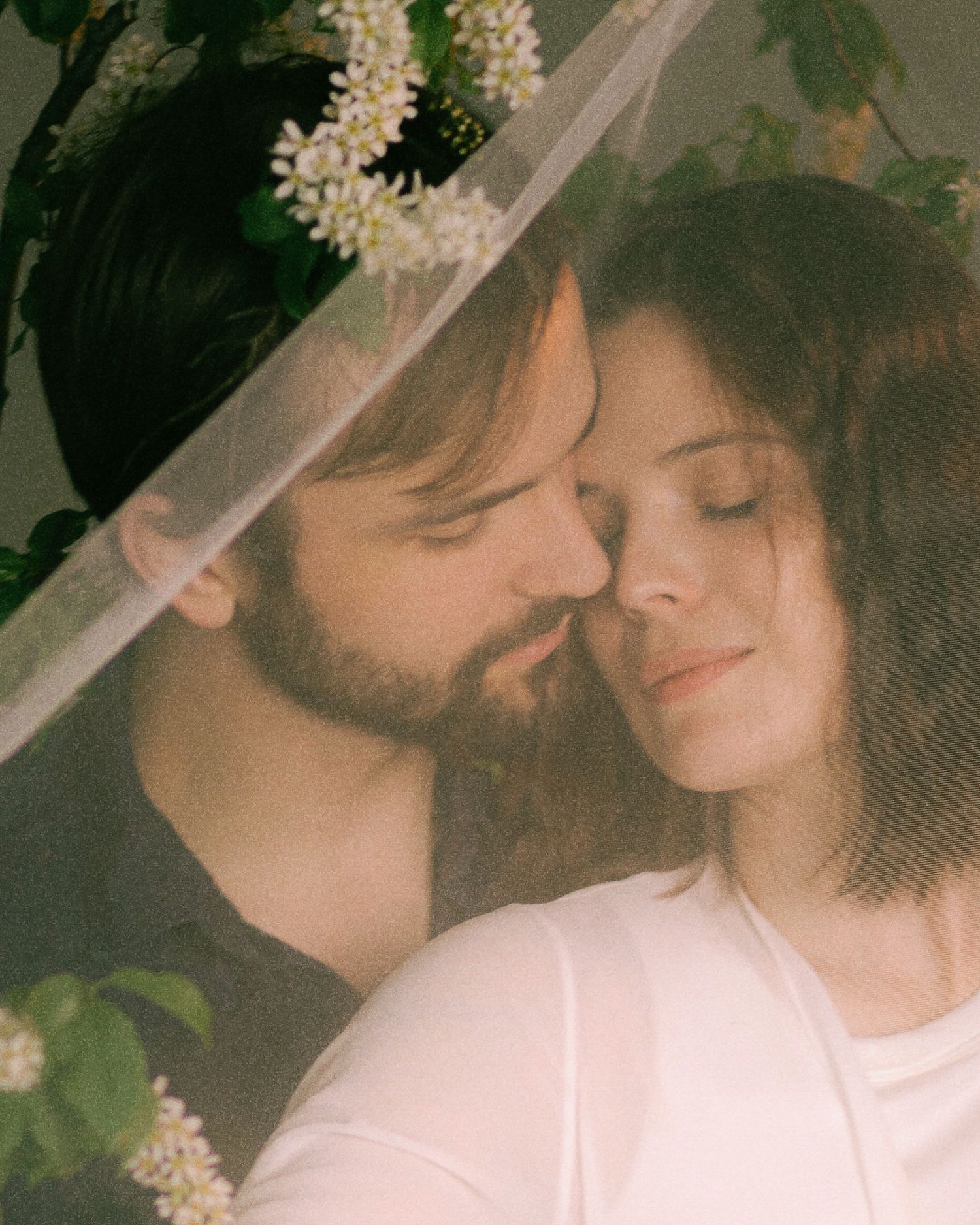 A romantic couple with closed eyes tenderly touching foreheads surrounded by white blooming flowers under a sheer veil.