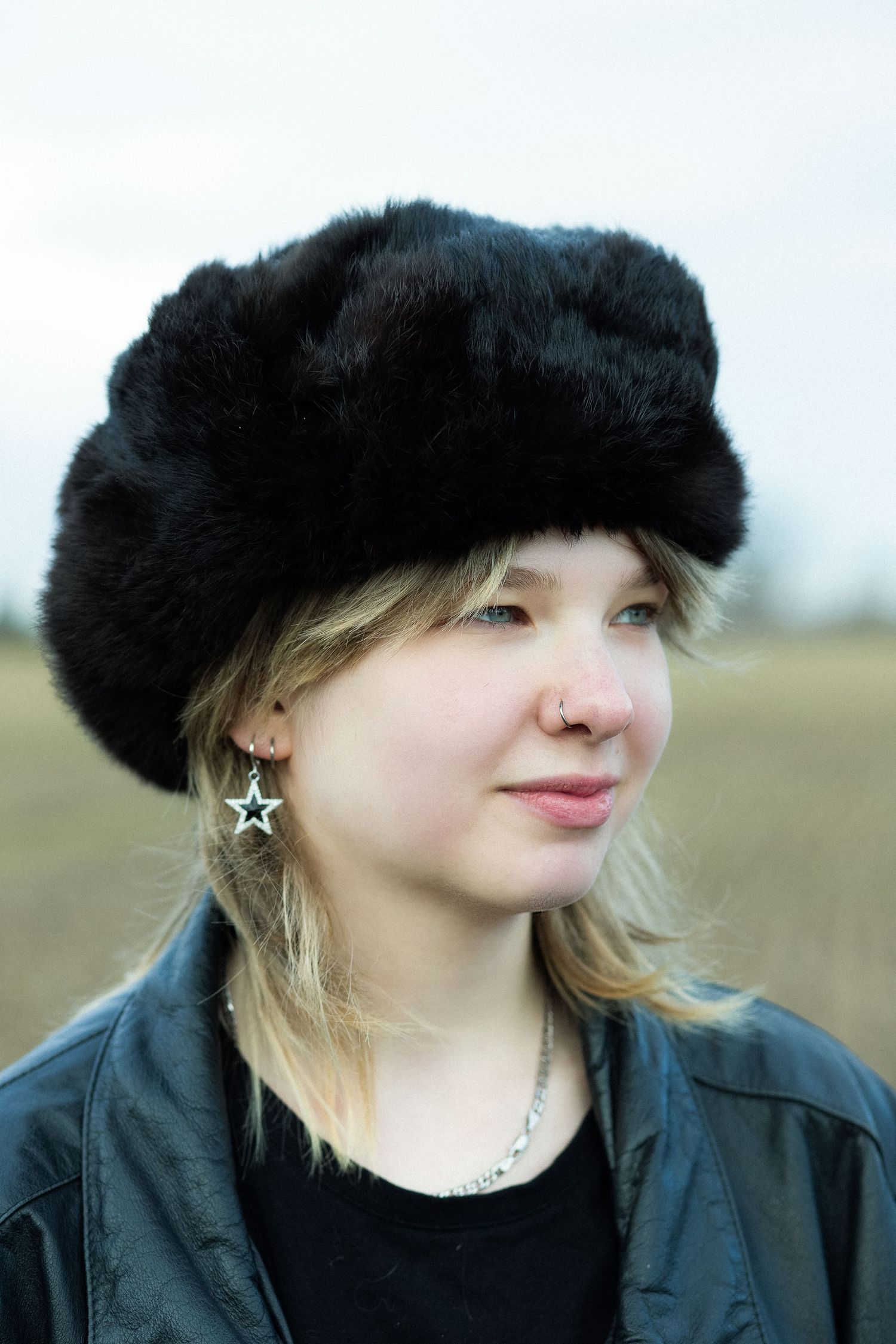 Young blonde woman wearing a black fur hat and leather jacket with star earrings and nose ring outdoors.