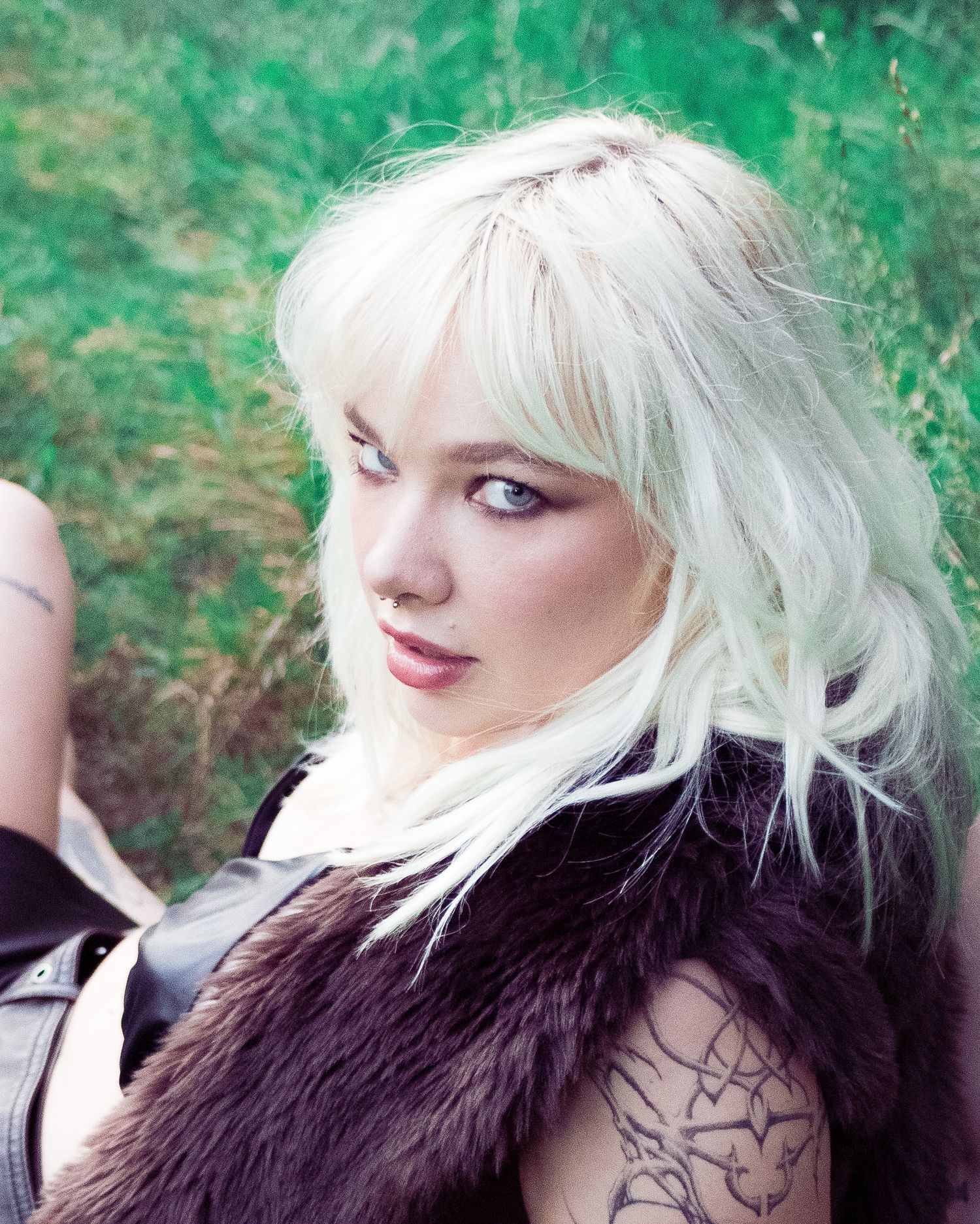 Platinum blonde woman with tattoos wearing a dark fur vest, looking over her shoulder in a lush green outdoor setting.