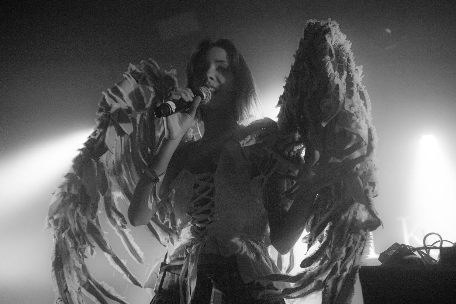 Black and white photo of a singer performing on stage wearing dramatic feathered wings costume under bright spotlights.