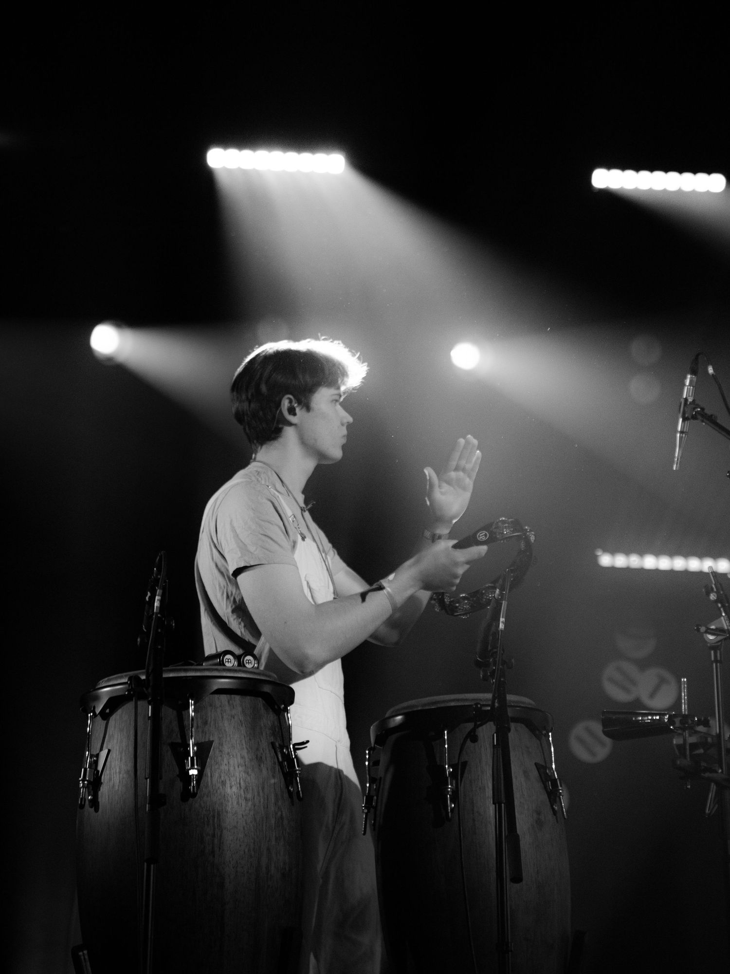 Black and white photo of a male percussionist playing congas under dramatic stage lighting at a live concert.