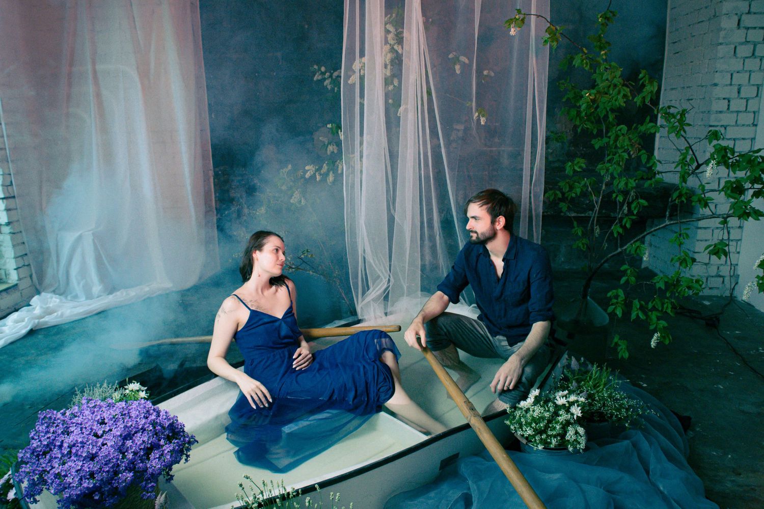 A pregnant woman in a blue gown sits in a bathtub with a bearded man, surrounded by lush plants and soft misty light.