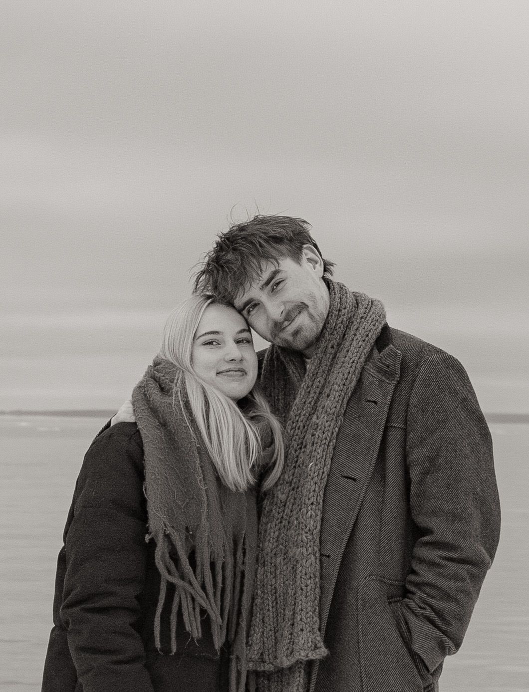 Black and white photo of a young couple in winter coats and scarves posing together outdoors near a beach.