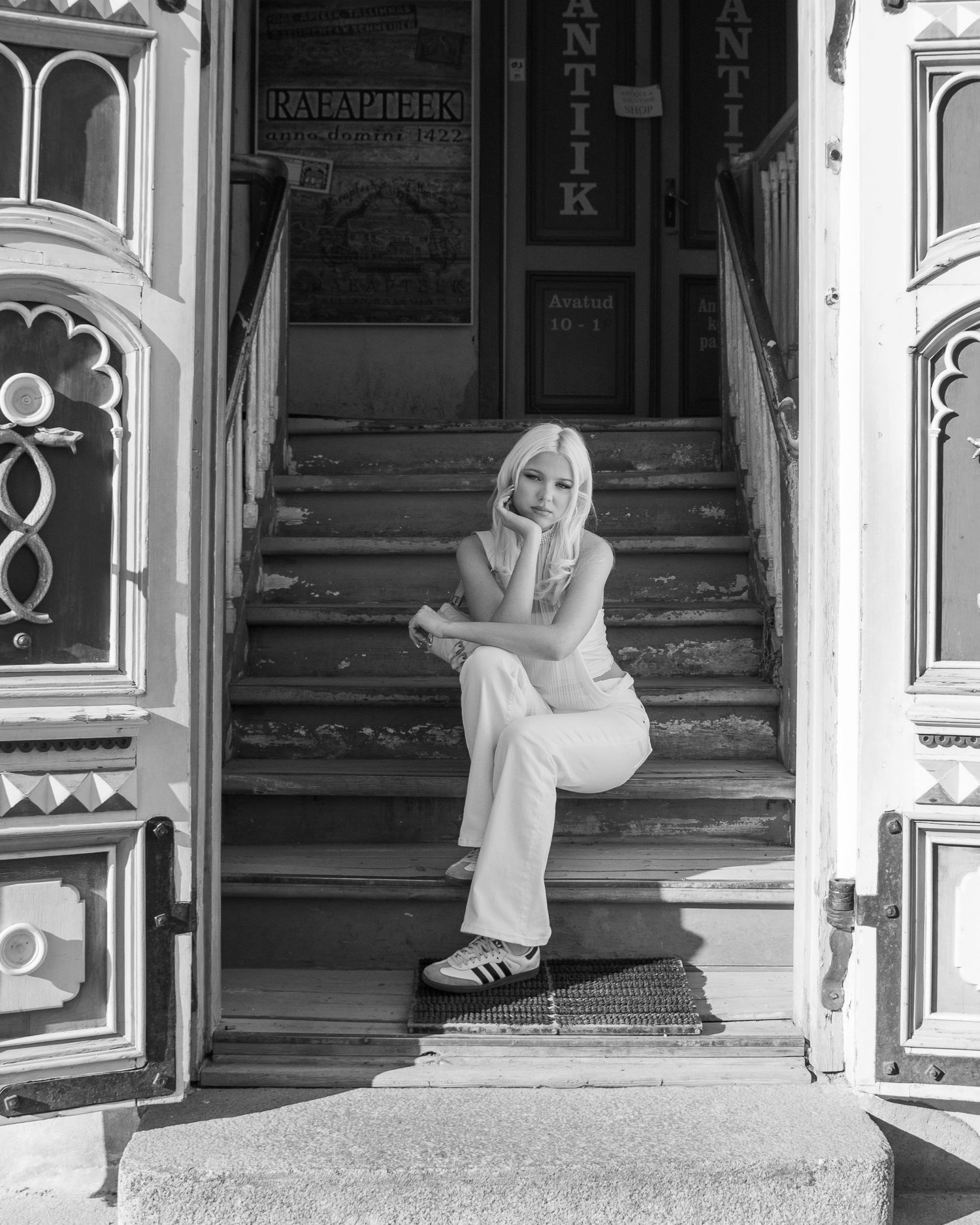 Black and white photo of a blonde woman sitting on vintage shop steps wearing white pants and Adidas sneakers.