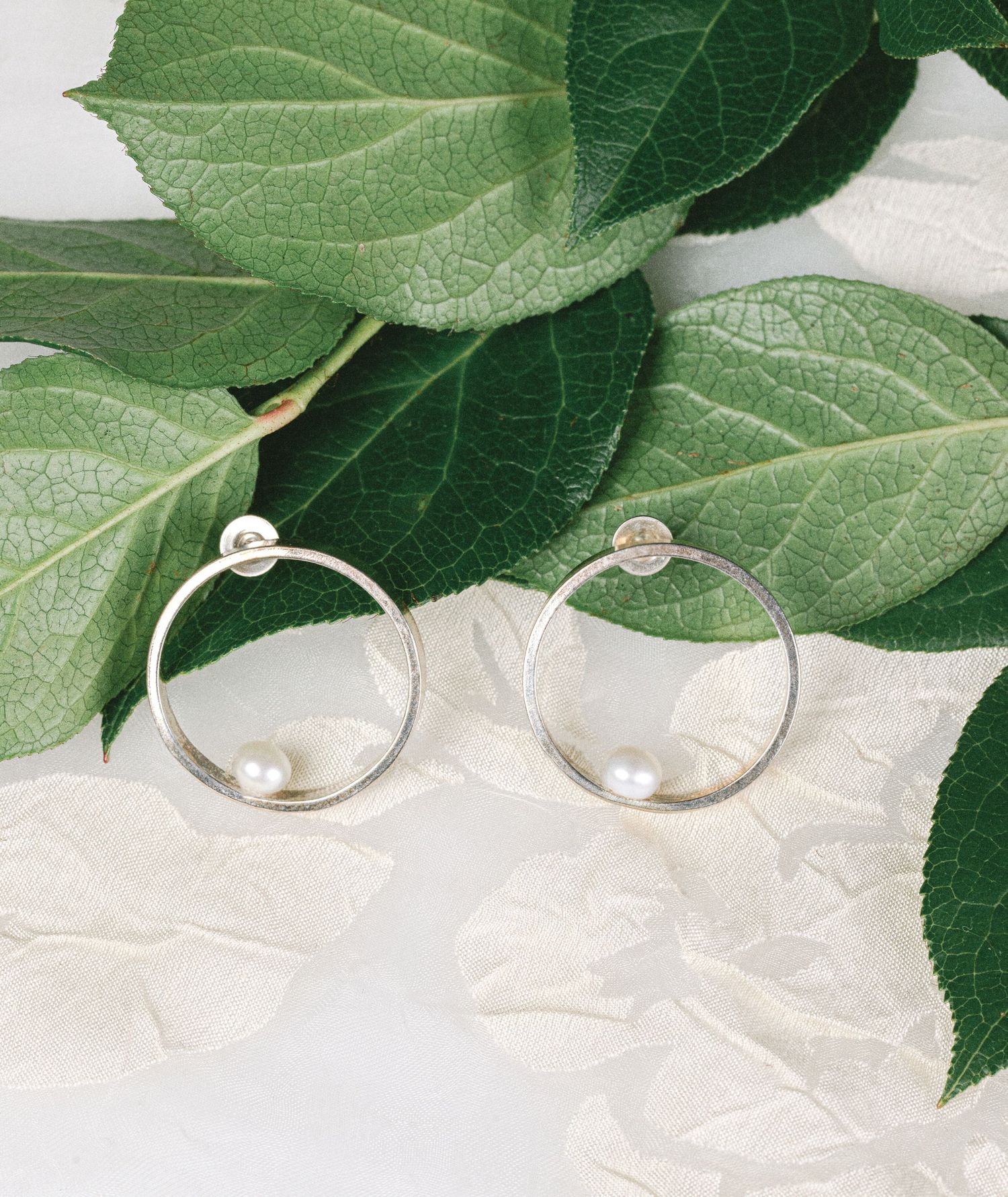Silver hoop earrings with freshwater pearl accents displayed on green leaves and white petals.