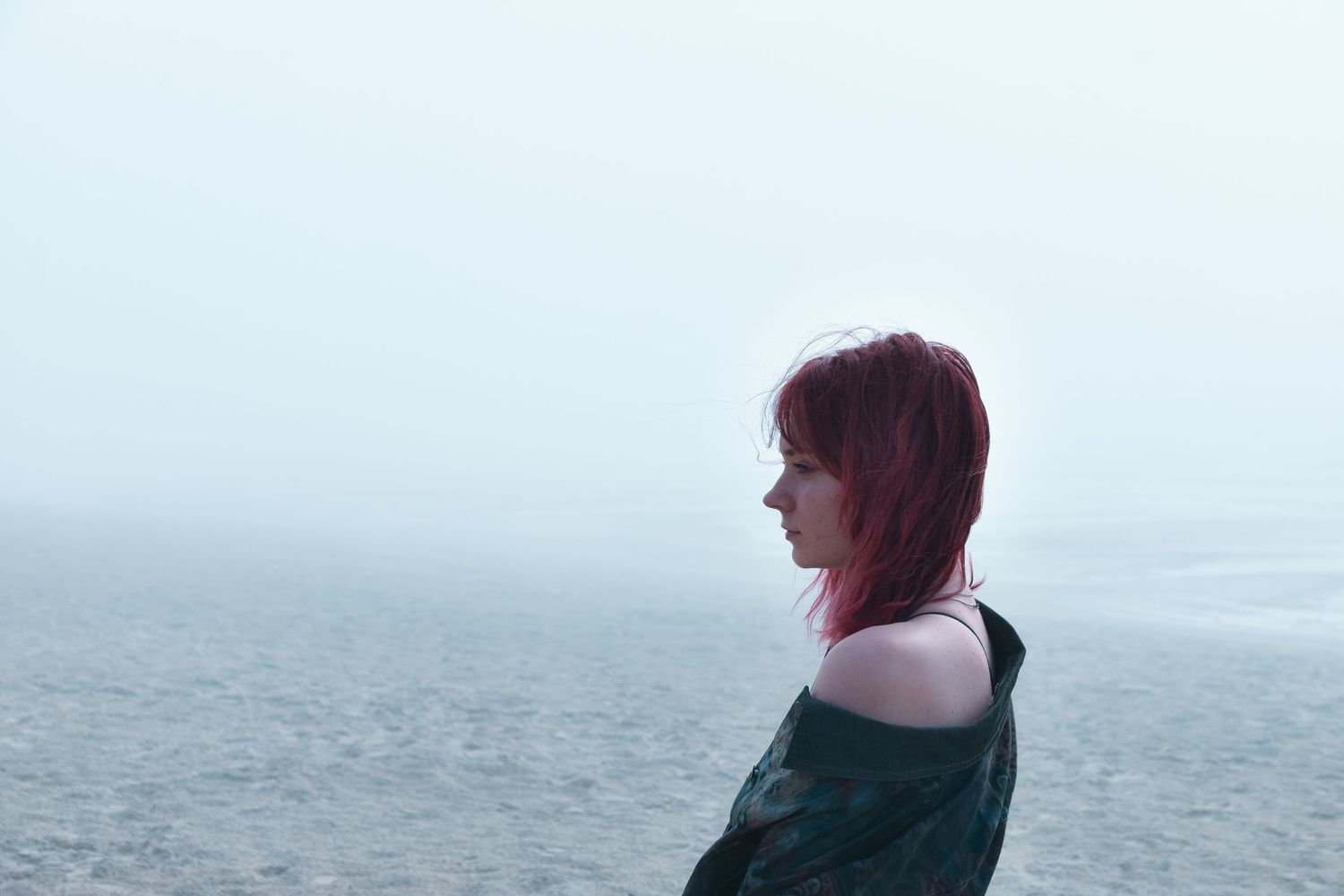 Side profile of a red-haired woman wrapped in dark fabric gazing over a misty ocean.