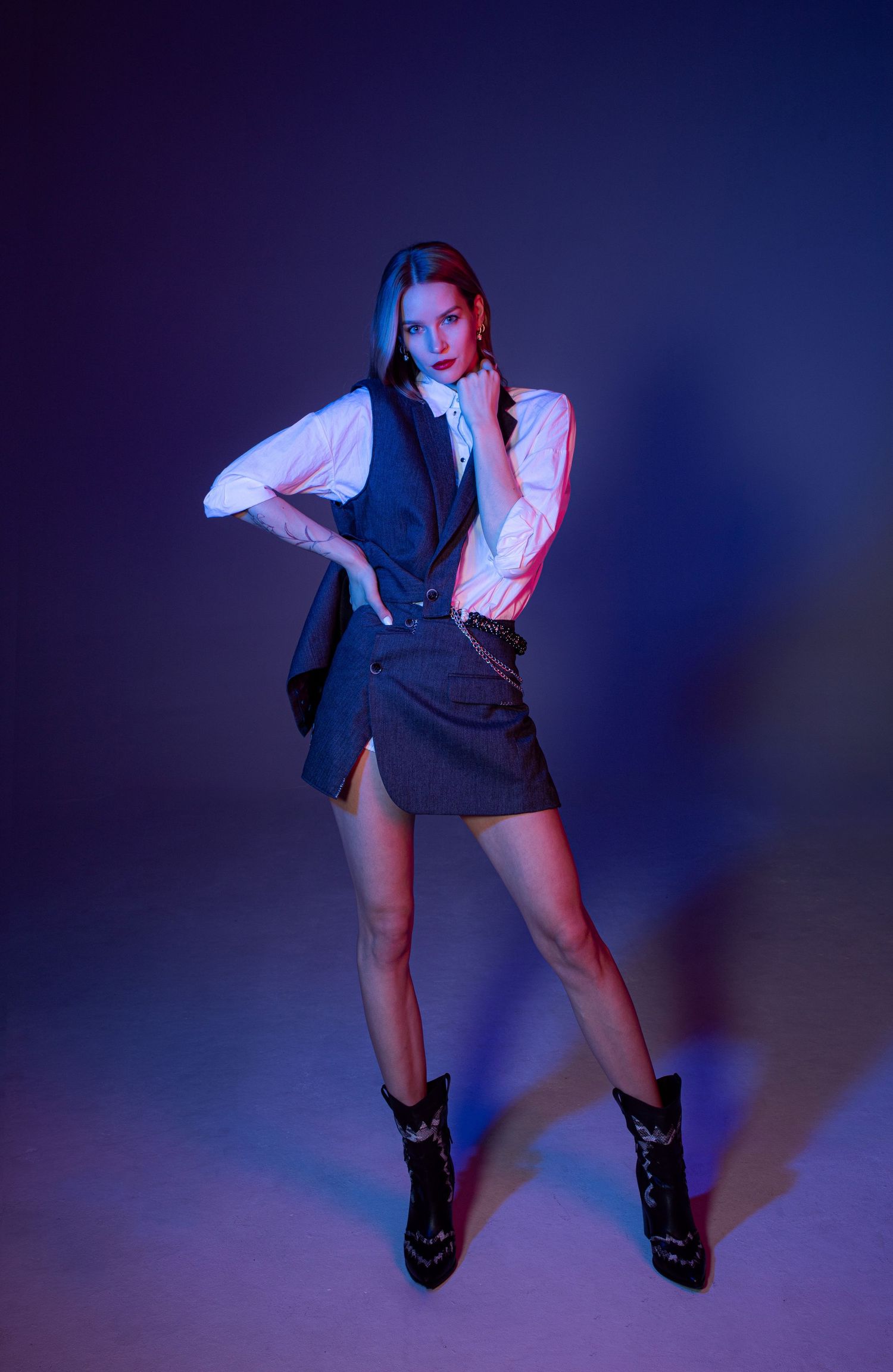 Brunette woman in white blouse and dark vest skirt poses boldly under blue and purple studio lights.
