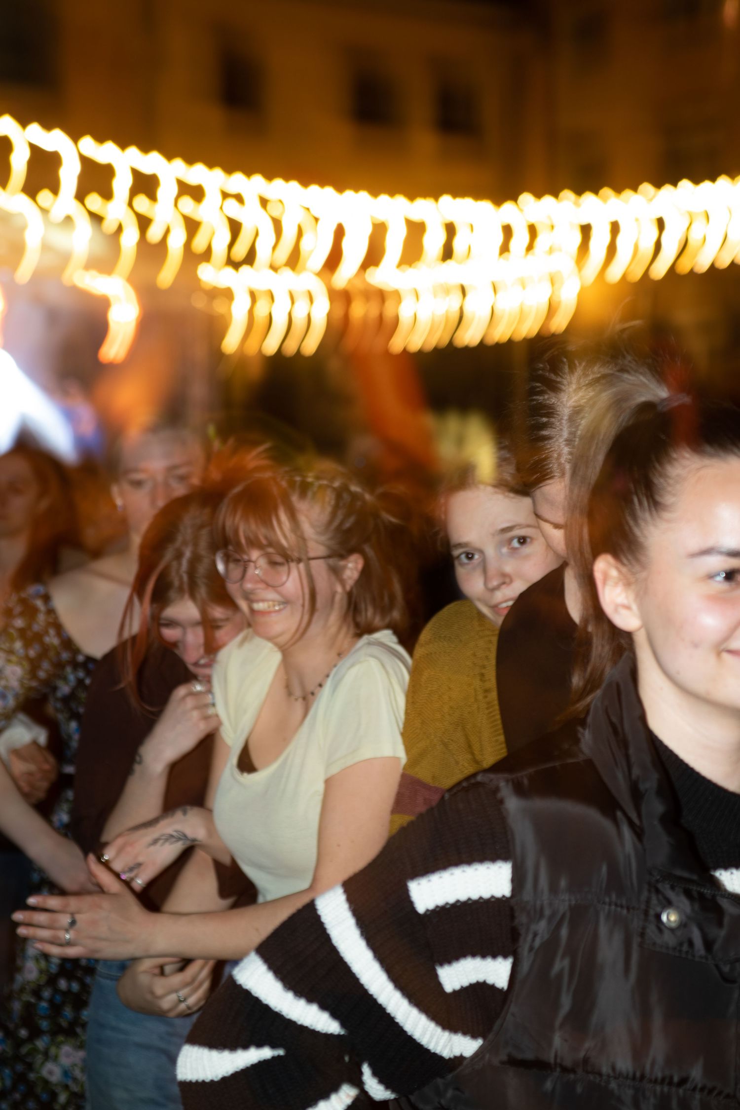 Group of young women dancing and smiling under warm string lights at an outdoor night event.
