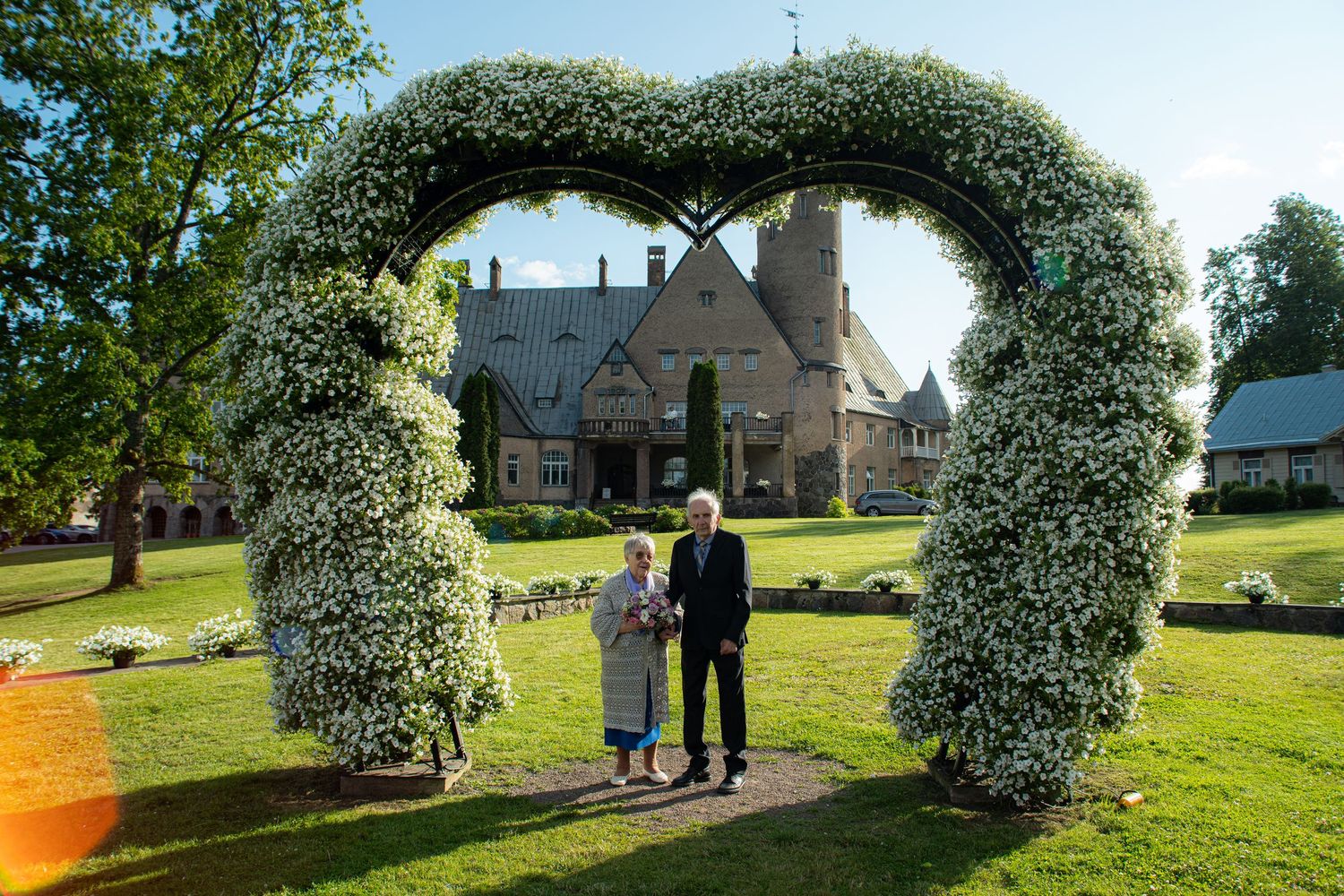Senior couple posing under a white floral heart arch in front of a grand historic stone estate.