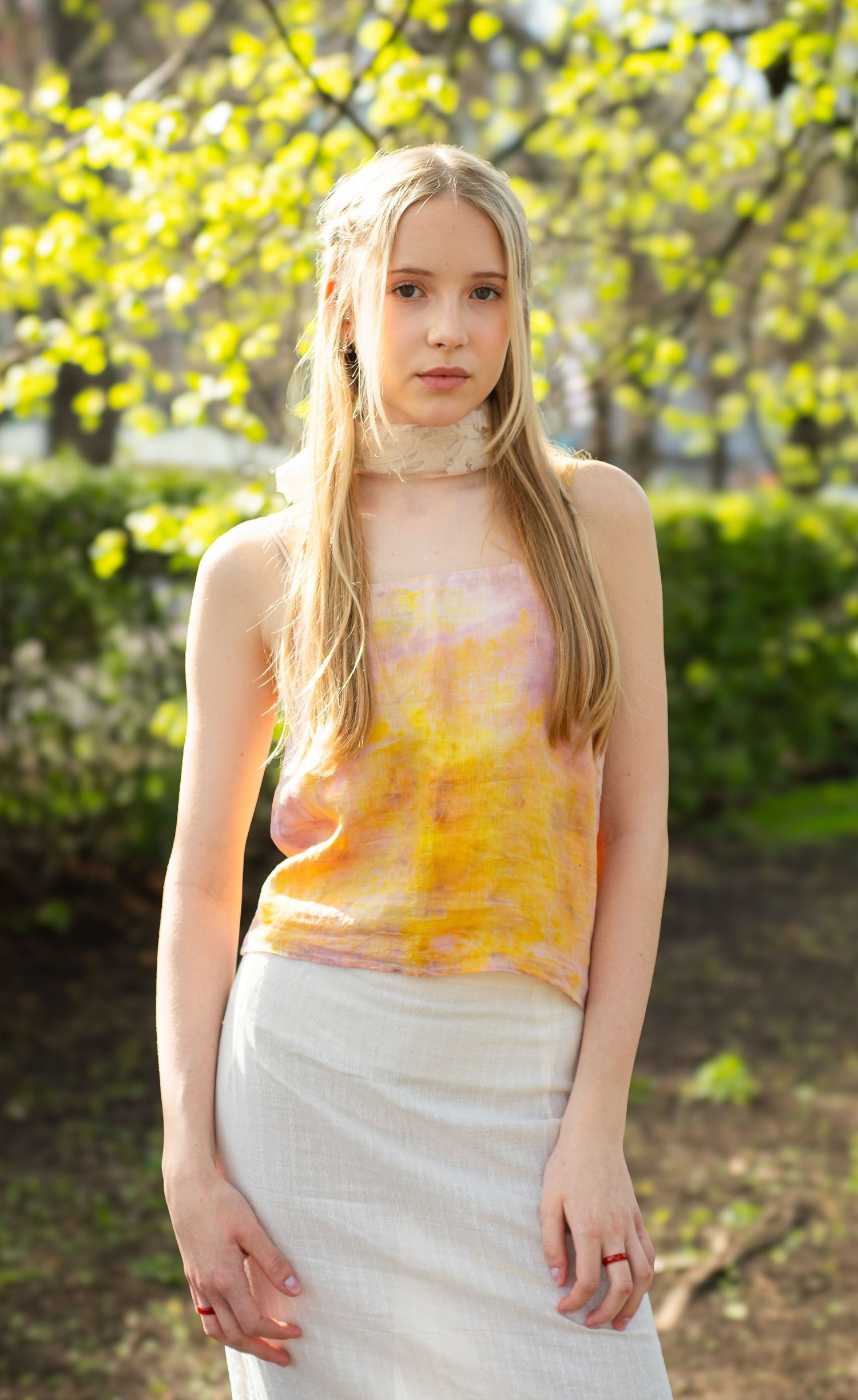 Young blonde woman in yellow tie-dye top and white skirt posing outdoors among spring trees.