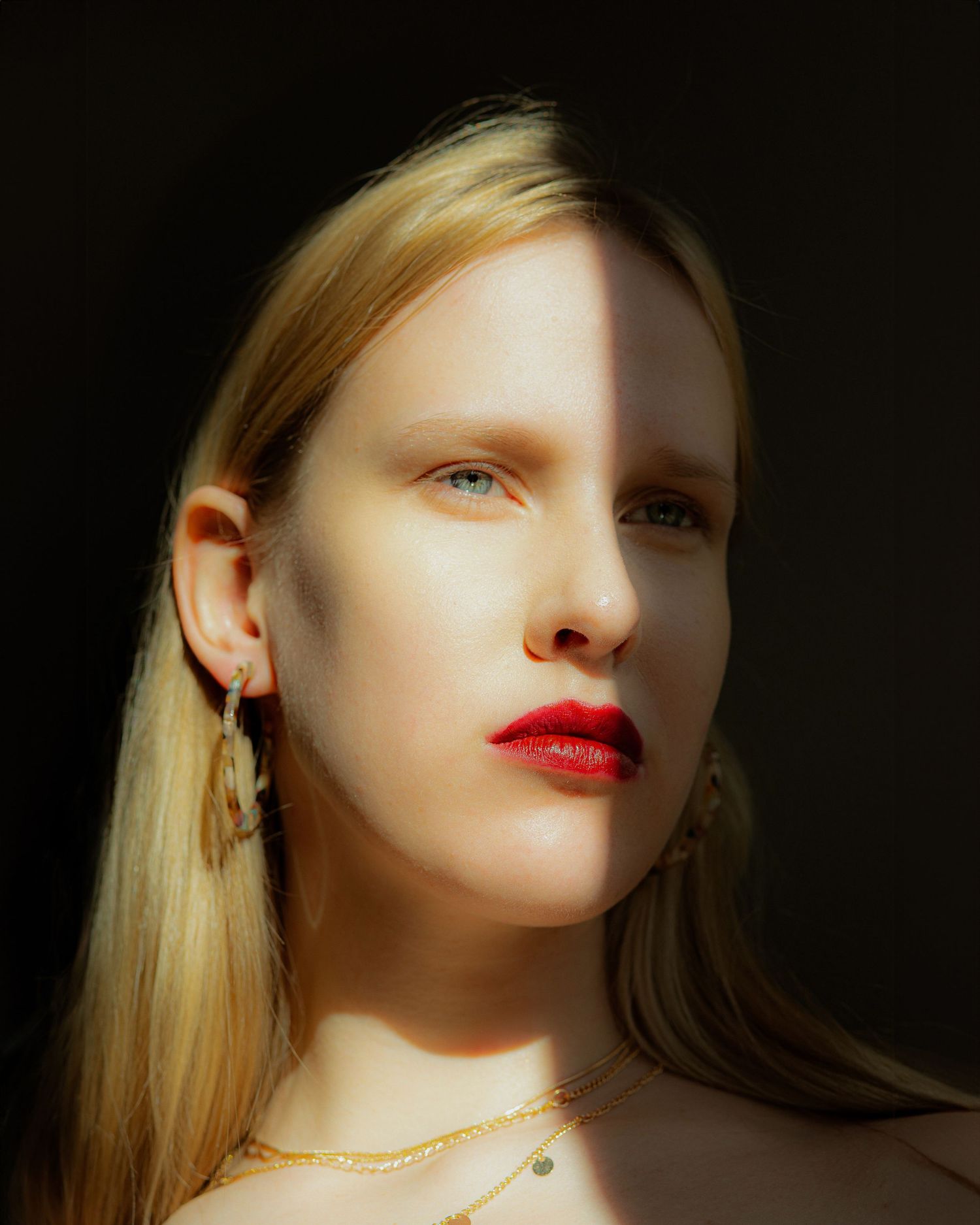 Blonde woman with red lips and gold jewelry photographed in dramatic half-shadow portrait.