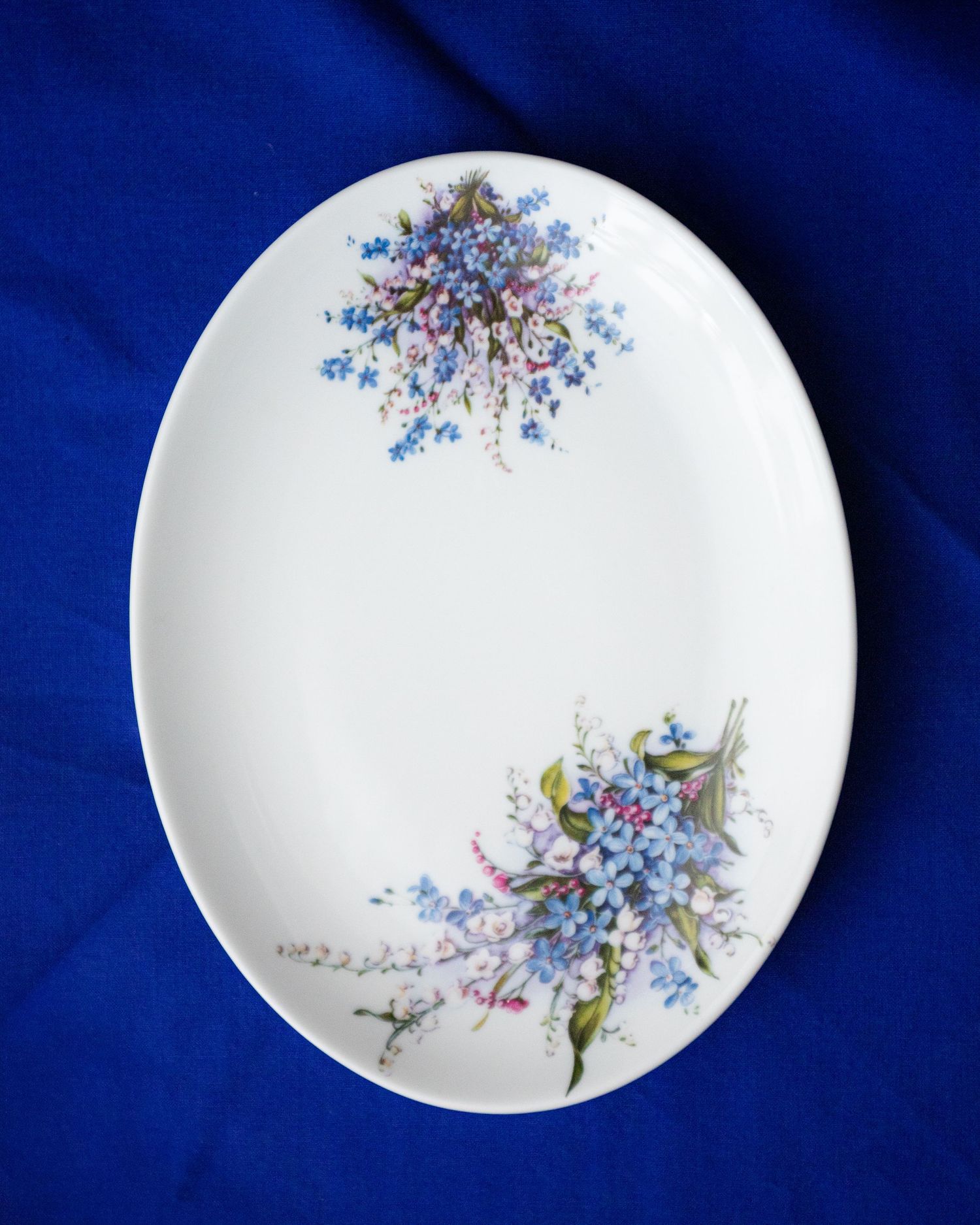 White oval porcelain plate with delicate blue forget-me-not floral bouquet designs on a royal blue background.