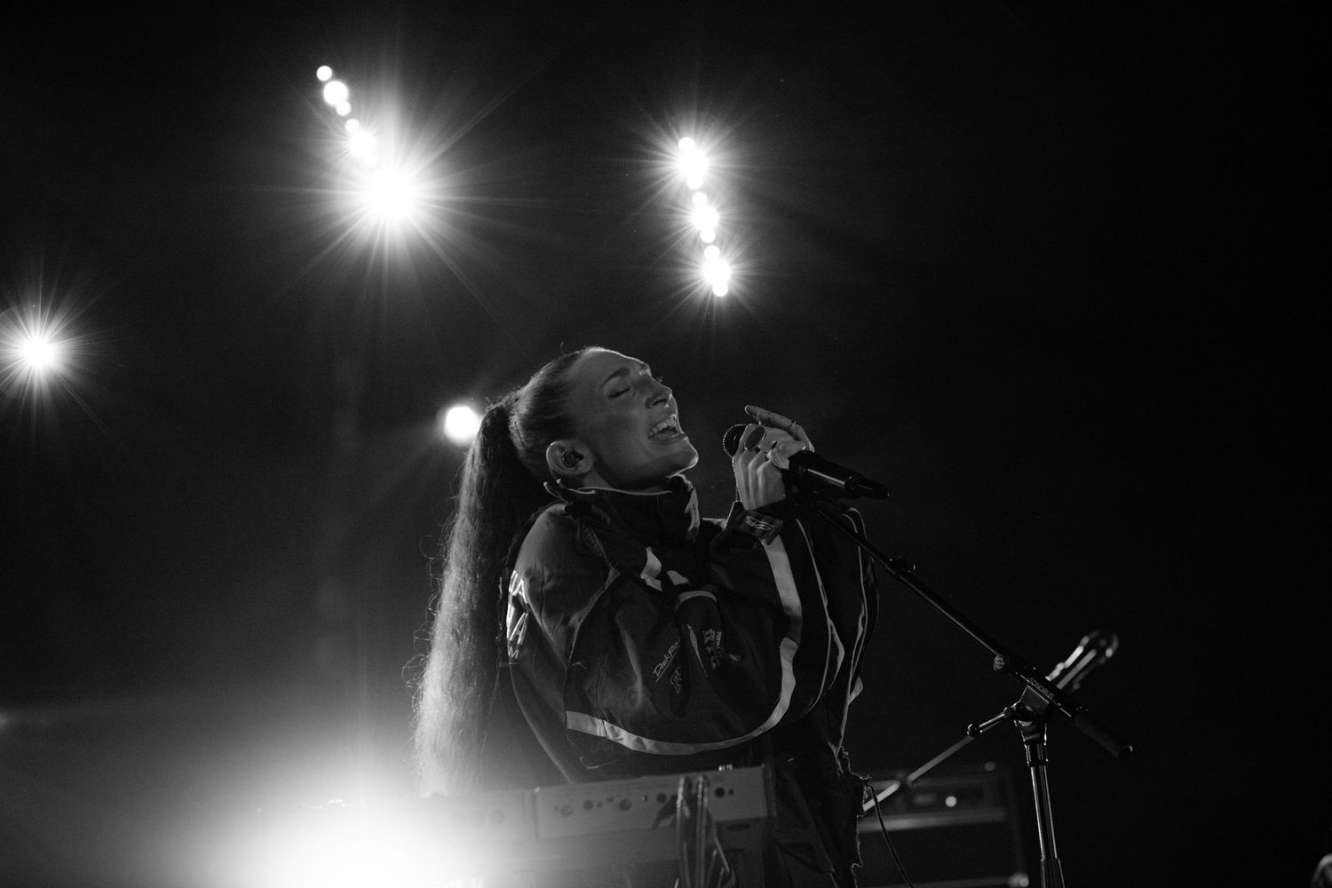 Black and white concert photo of singer performing at microphone under bright stage spotlights.