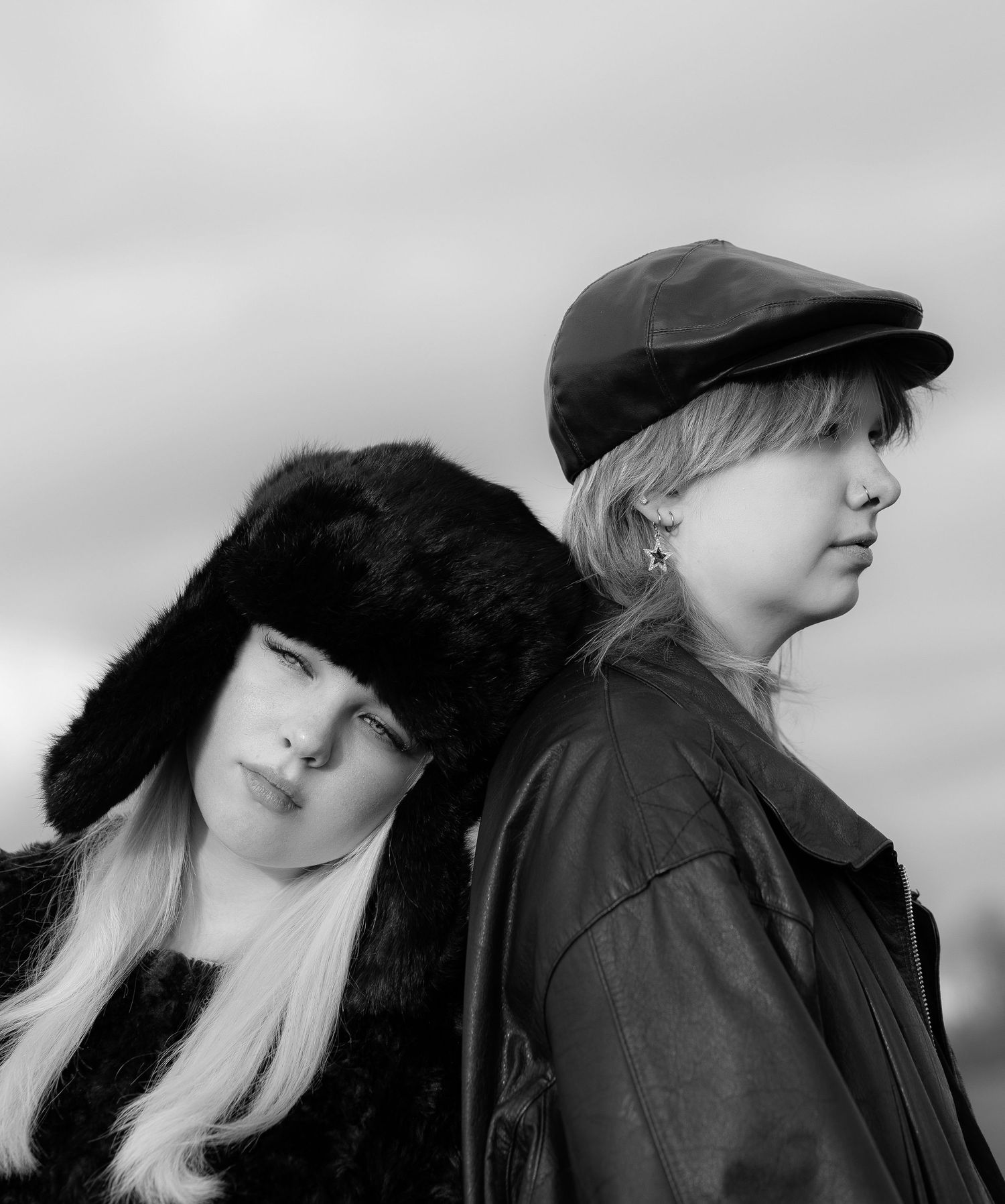Black and white portrait of two young women wearing fur and leather hats against cloudy sky.