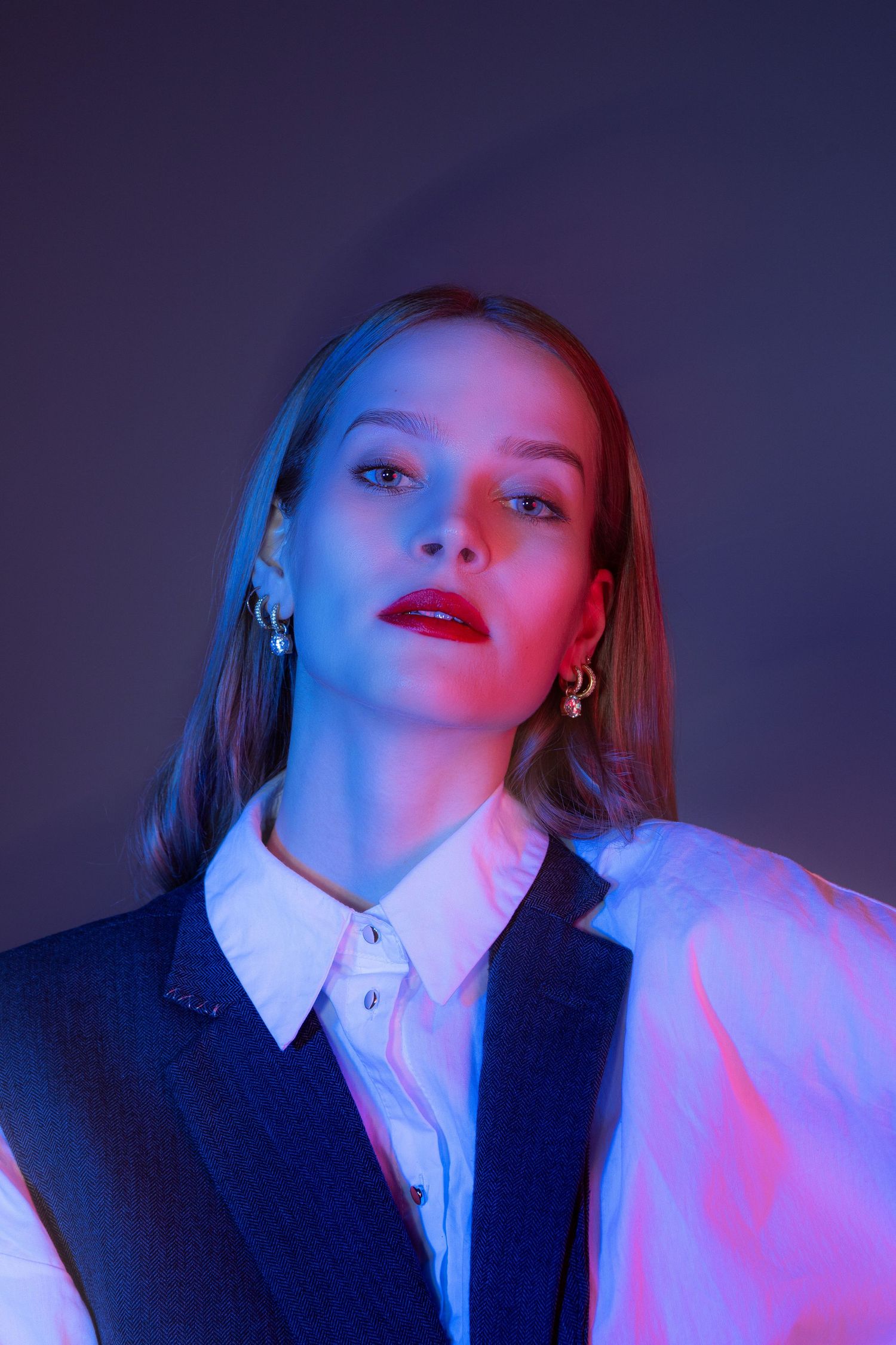 Portrait of woman in dark vest and white shirt with red lips under blue and pink studio lighting.