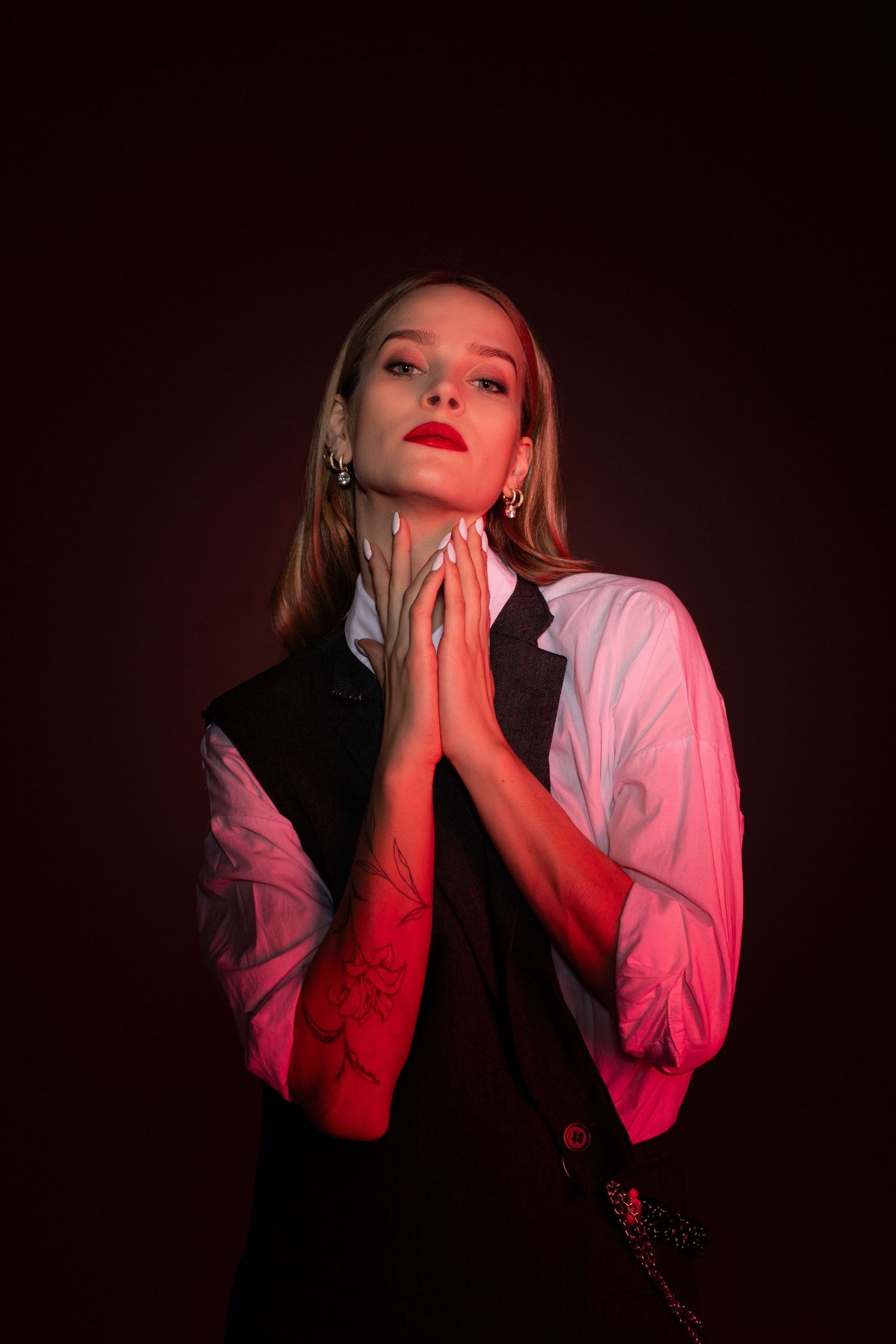 Woman with red lips in prayer pose wearing white shirt and black vest under dramatic red lighting.